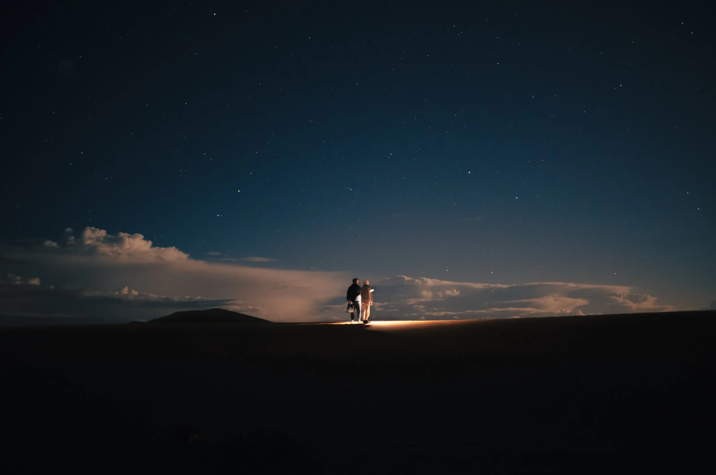 Two people stand together at night on a hill under a star-filled sky, with clouds and distant mountains in the background.