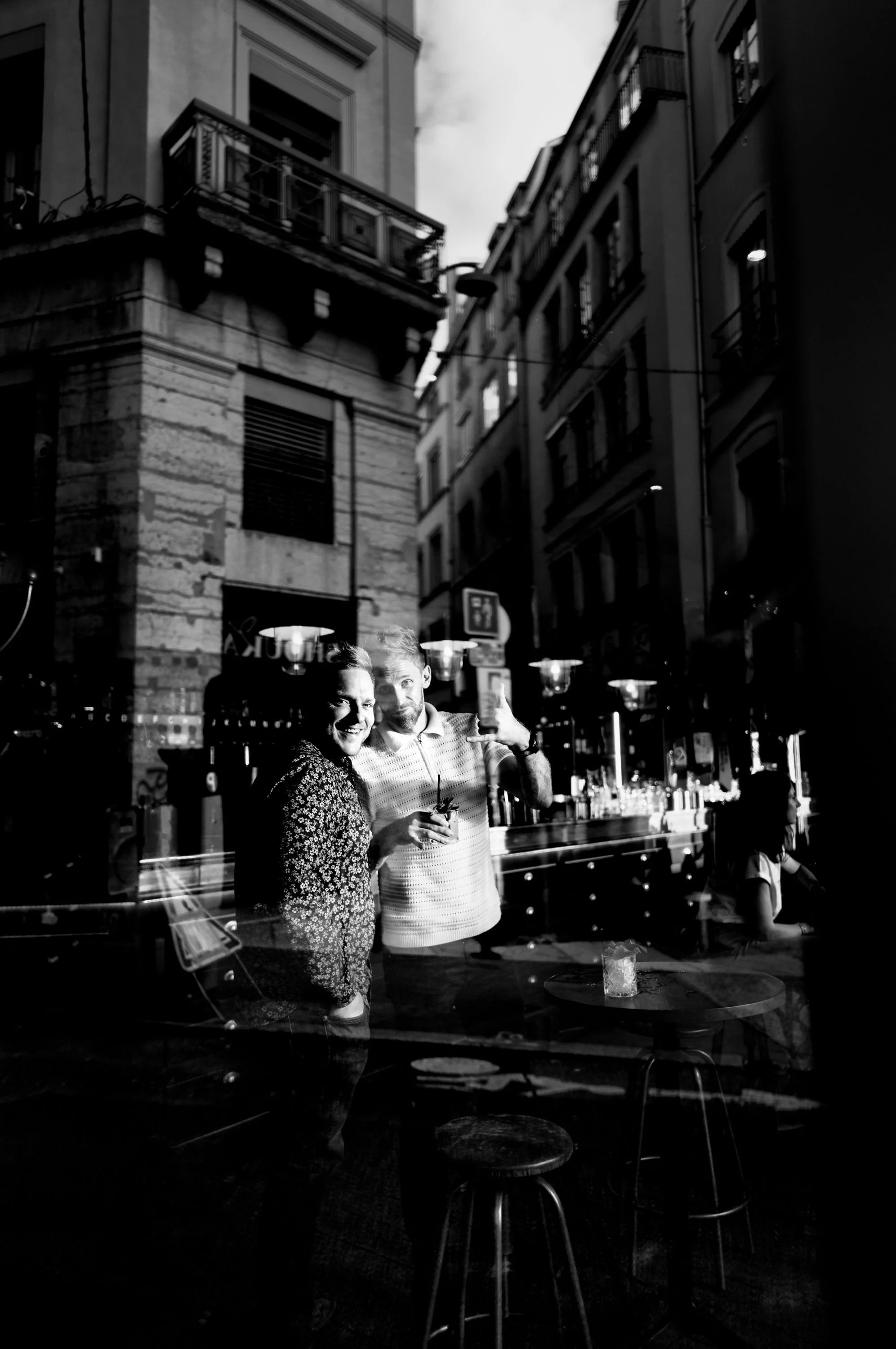 Reflection of a smiling man and woman taking a selfie inside a bar, with city buildings visible outside through the window.