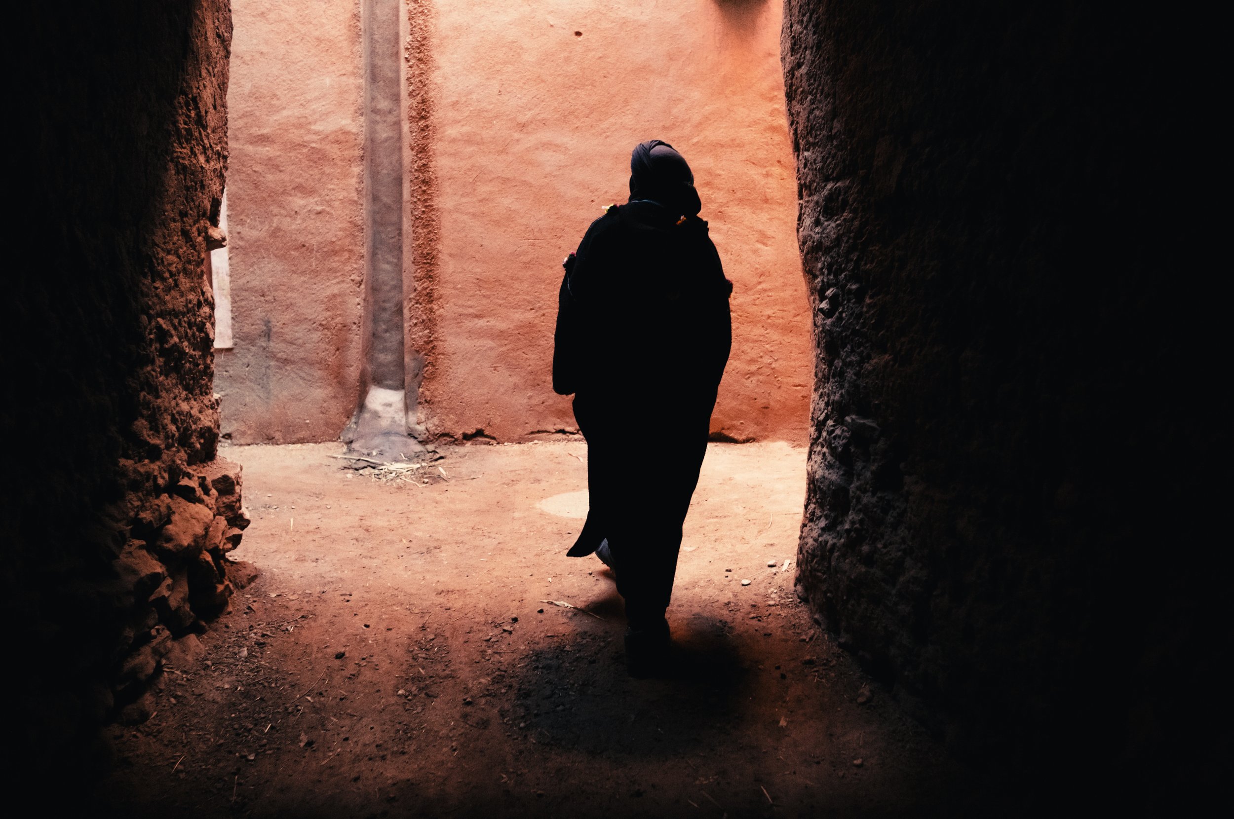 Silhouette of a person walking through a narrow passage between two brown mud walls, with bright light at the end of the passage.