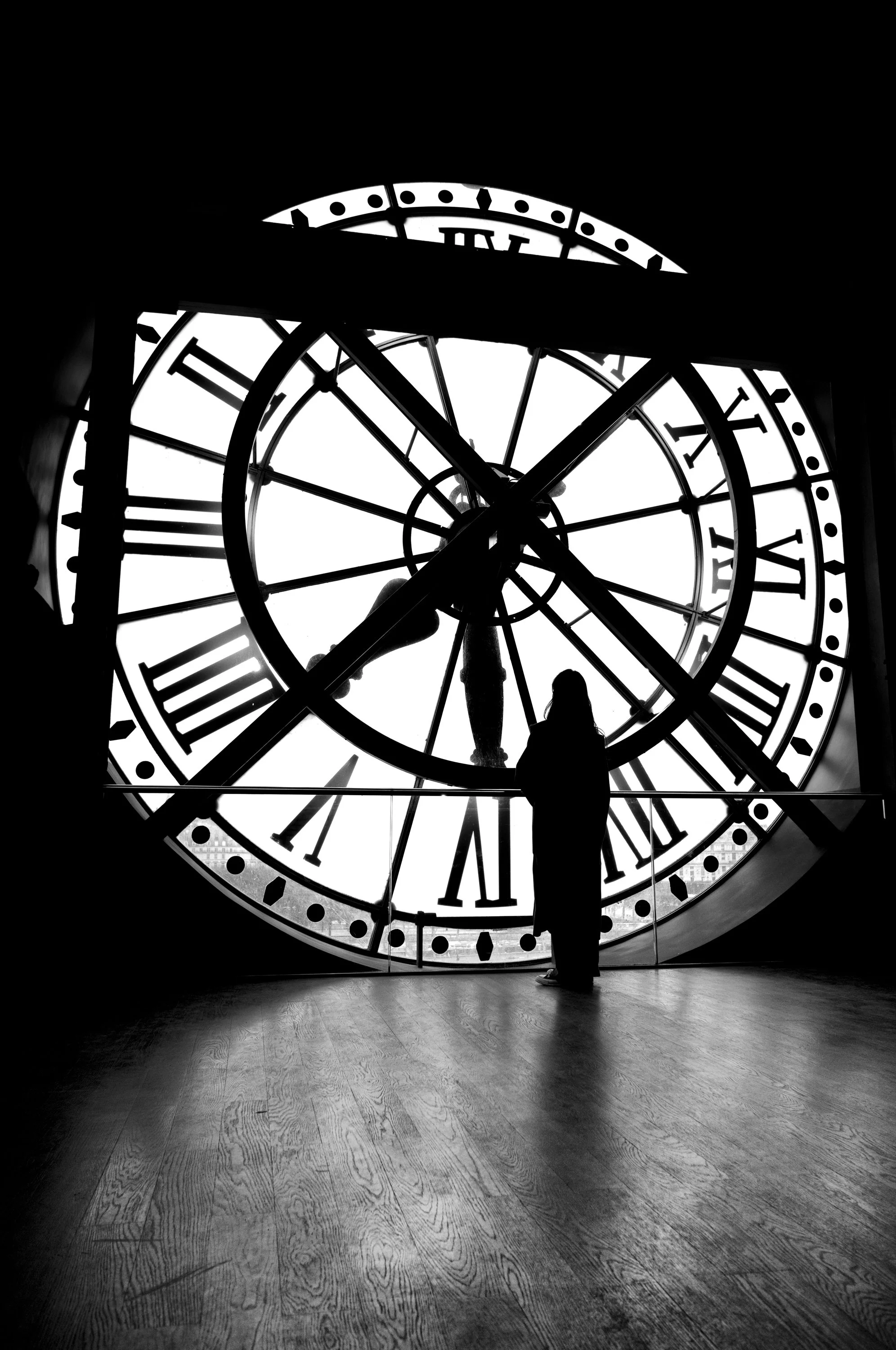 Silhouette of a person standing inside a clock tower, looking out through a large glass clock face showing Roman numerals, with a wooden floor in the foreground.