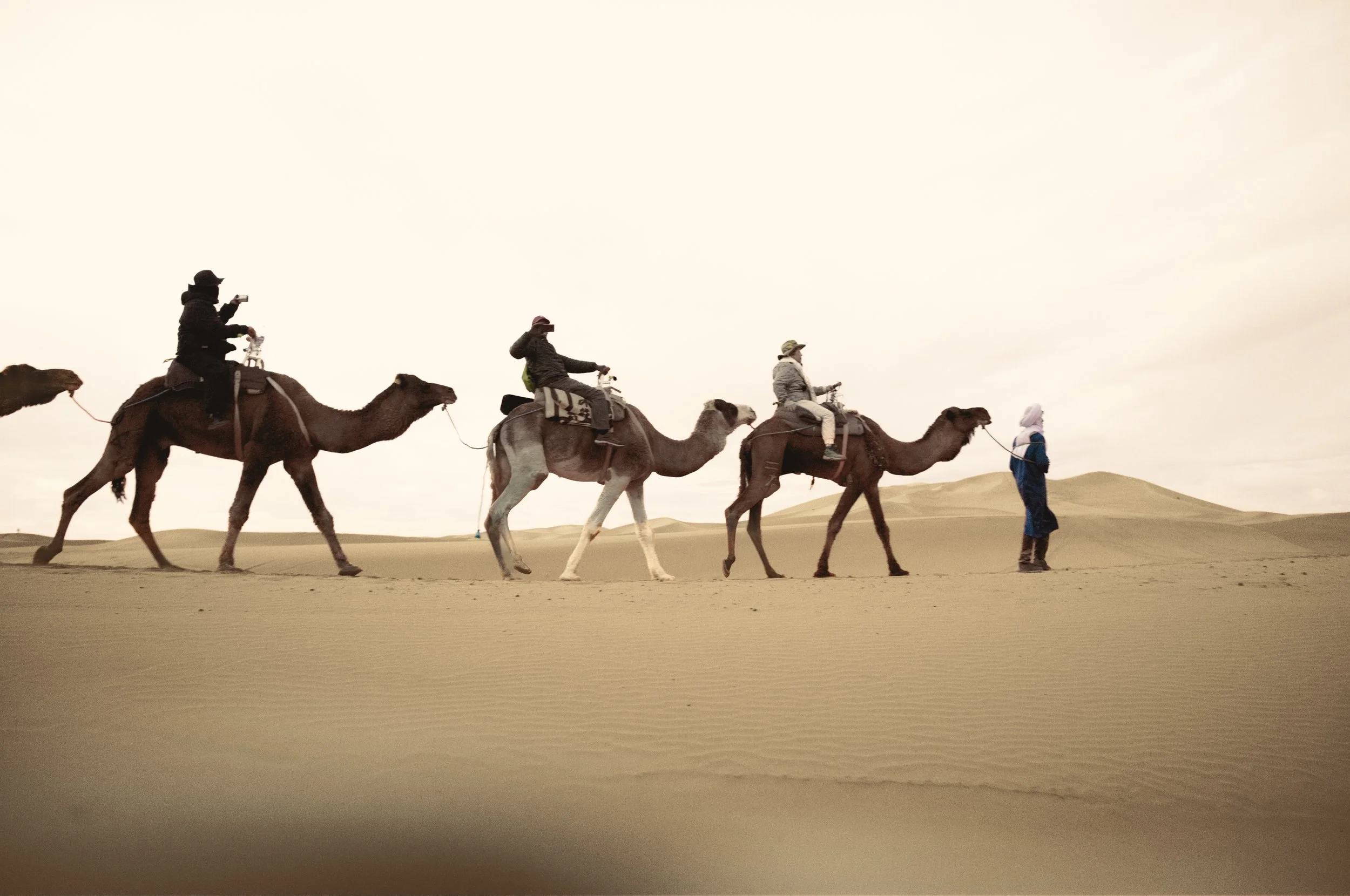People riding camels in a desert, with a guide walking alongside.