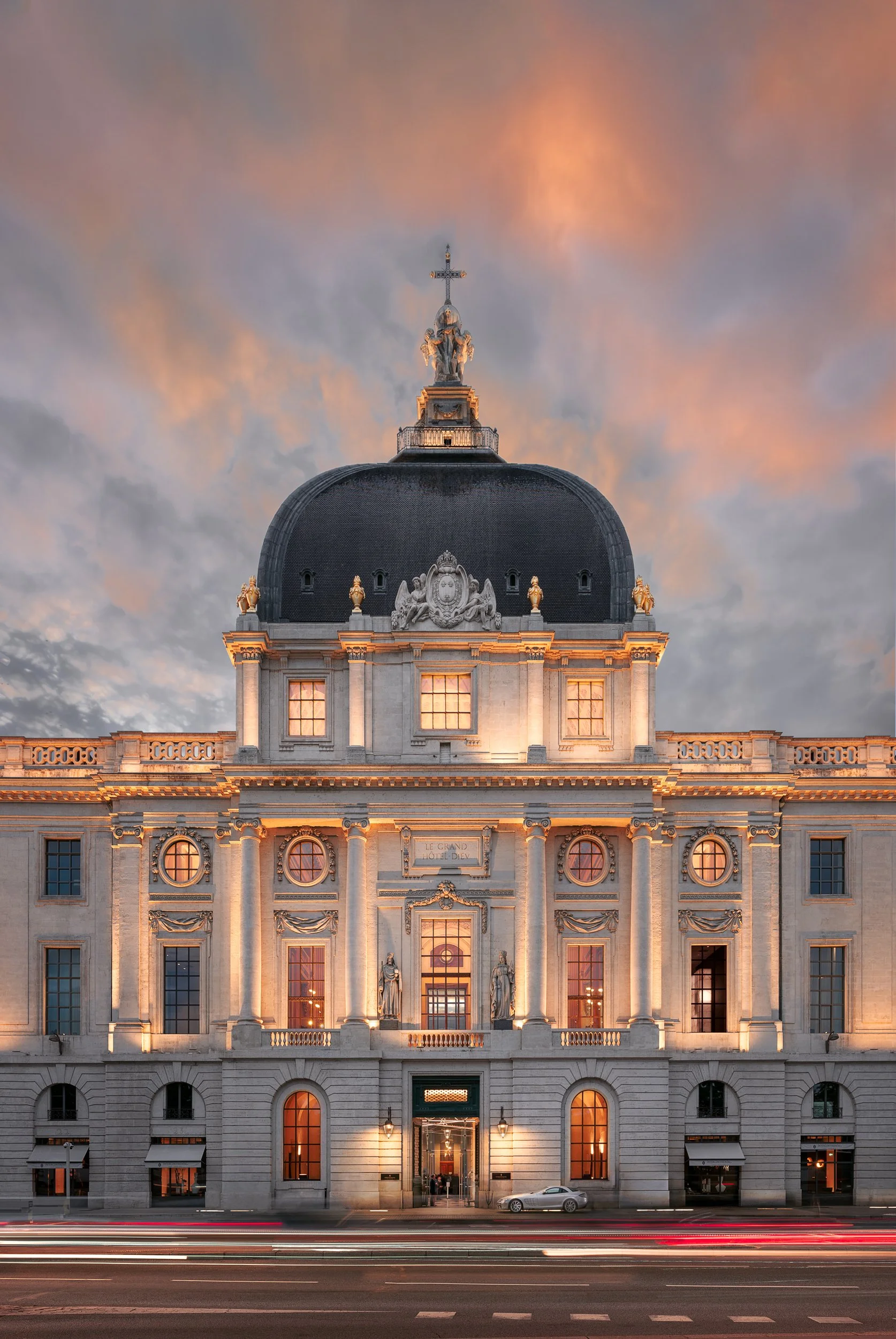 A historic building with ornate architecture, illuminated at dusk, with a dramatic cloudy sky in the background and streaks of car lights in motion at the street level.