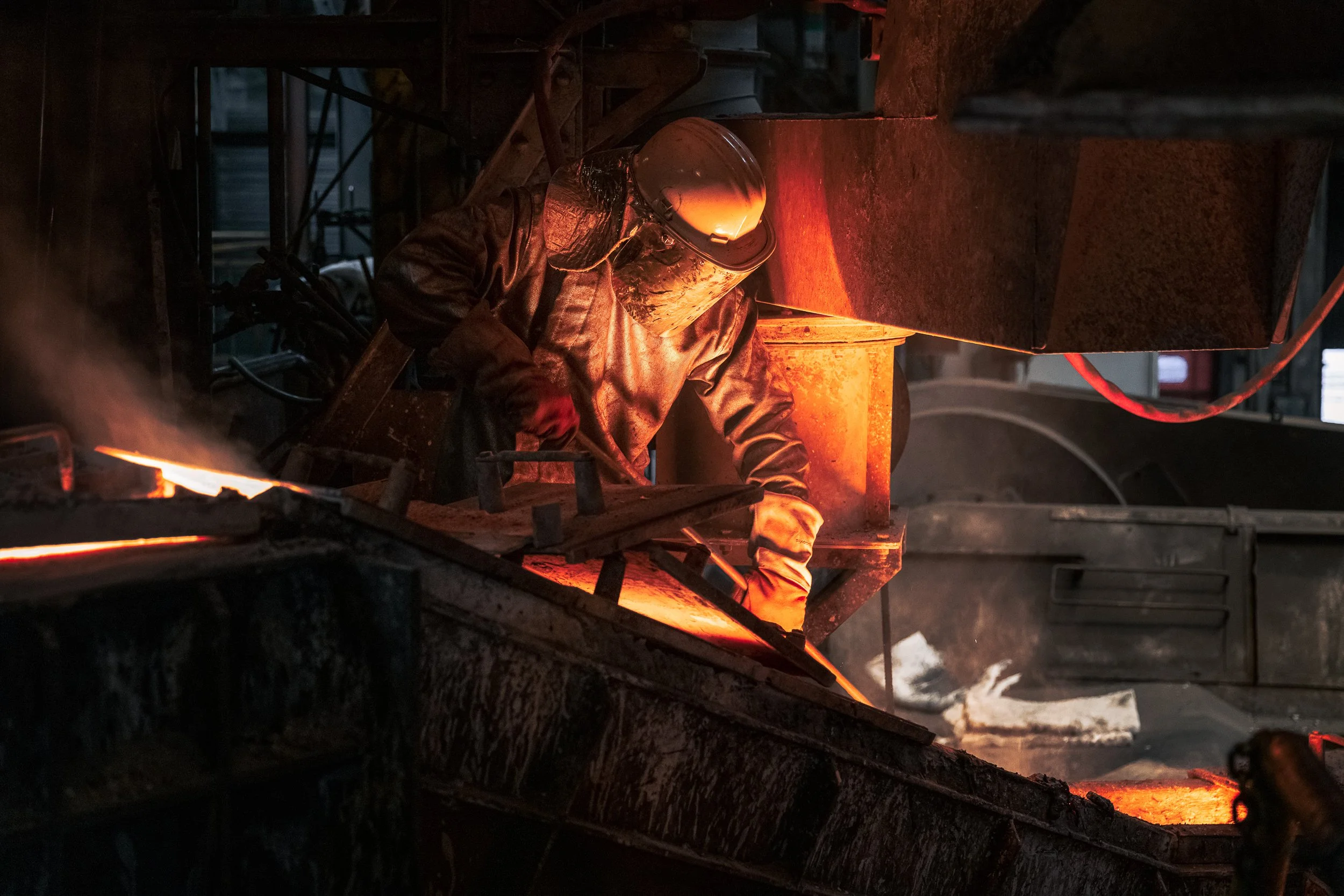 A worker in protective gear, including a welding helmet and gloves, is working on molten metal in an industrial foundry with glowing hot surfaces and machinery around.