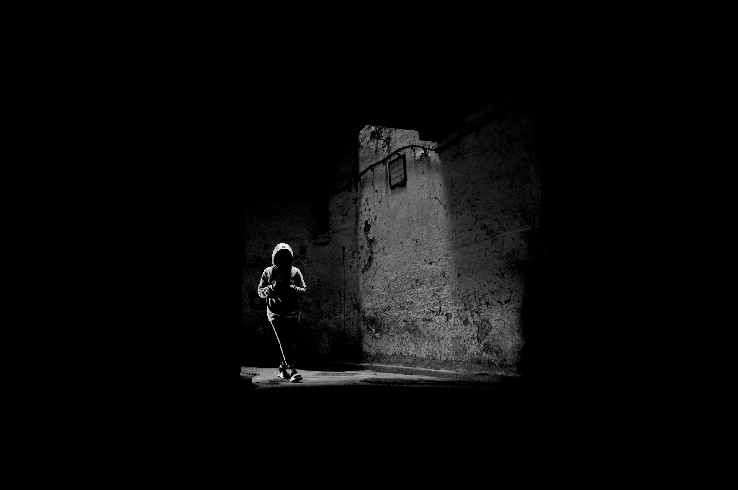 Child with a hoodie walking with a stick in a dark alley, illuminated by a small beam of light on weathered wall.