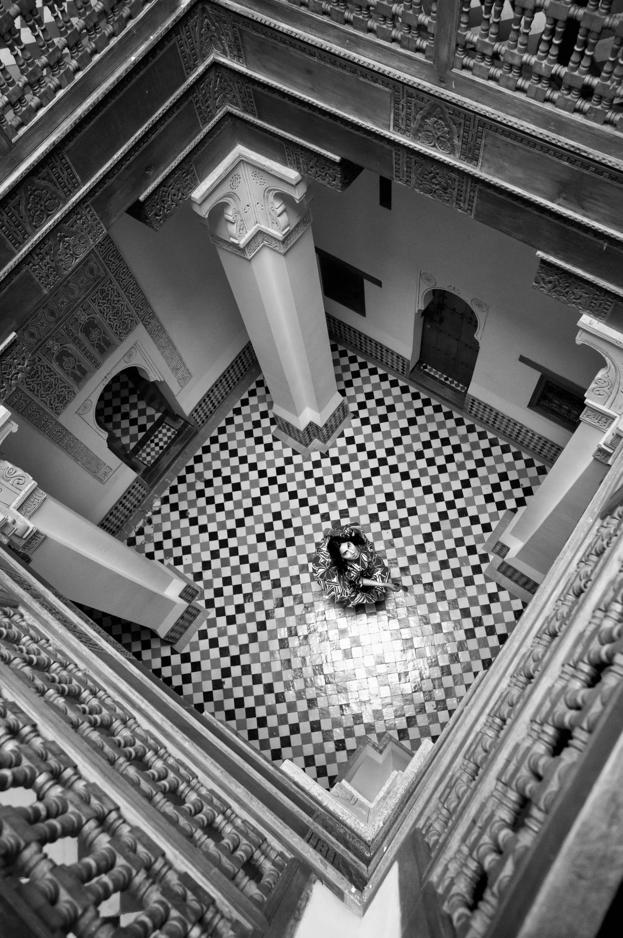 A woman looking up from the center of an ornate, multi-level staircase with intricate woodwork and a black-and-white checkered floor.