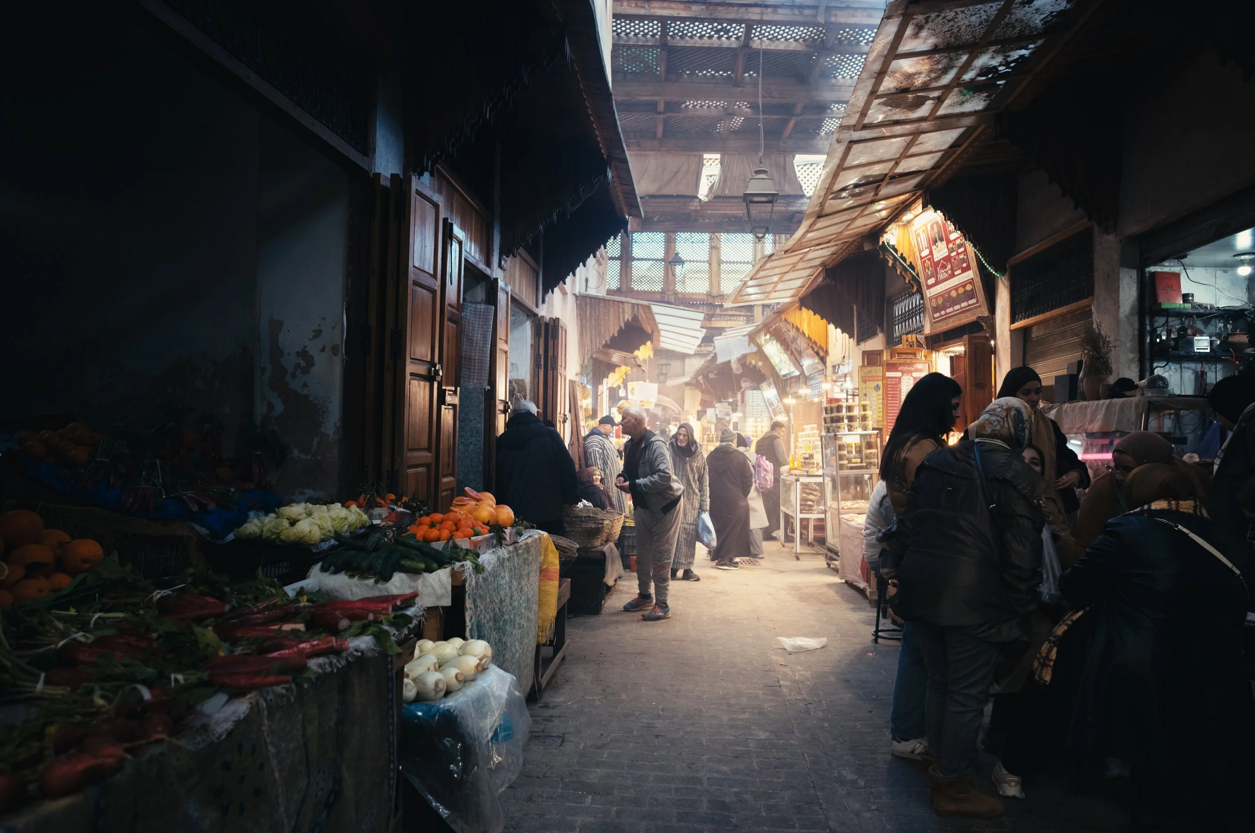 A bustling indoor market with vendors selling fresh vegetables and fruits on stalls, and customers shopping and socializing inside a covered space with wooden beams and diffused sunlight.