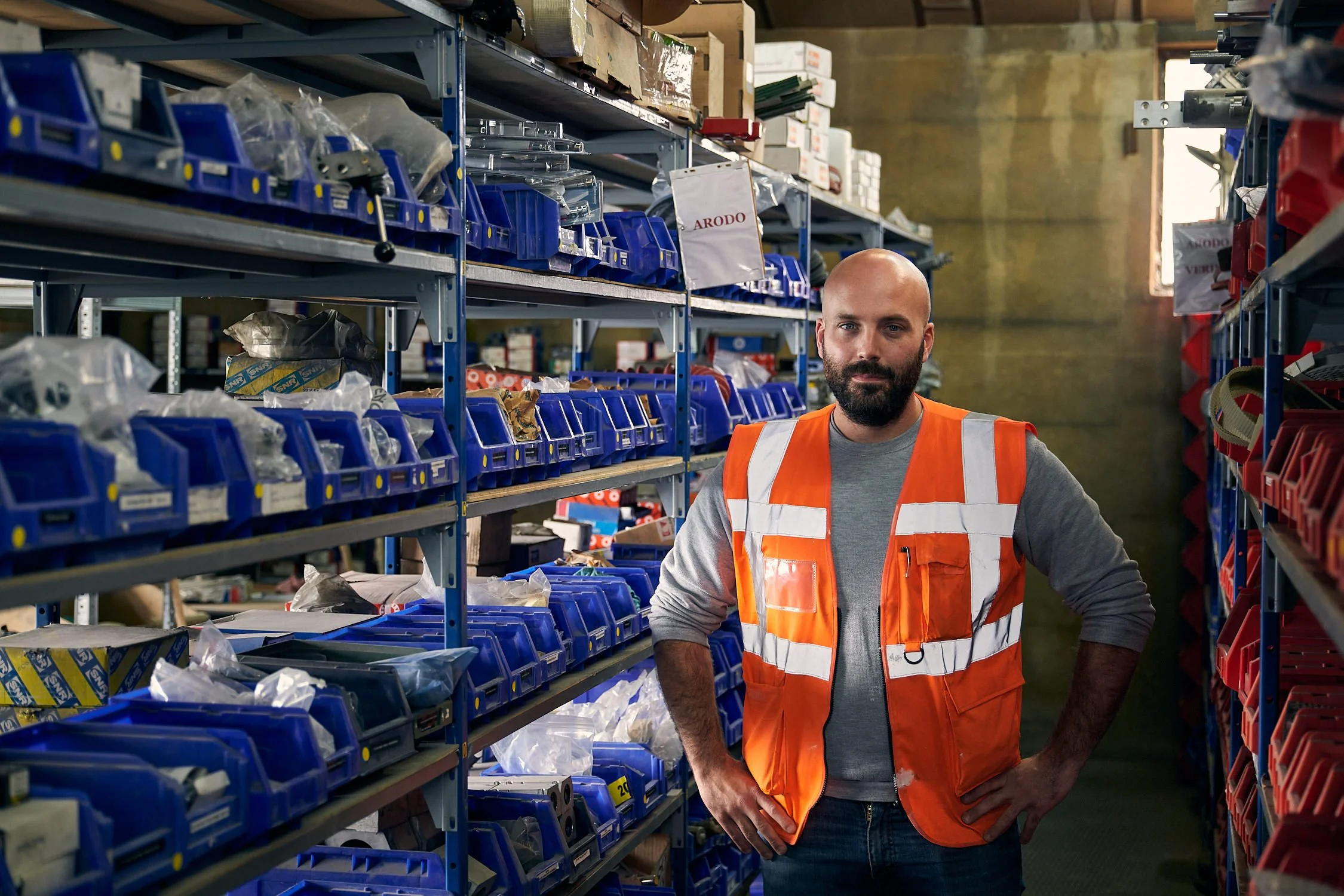 A man with a beard wearing an orange safety vest and gray long sleeve shirt standing with hands on hips in a warehouse aisle filled with blue and red storage bins on metal shelves.