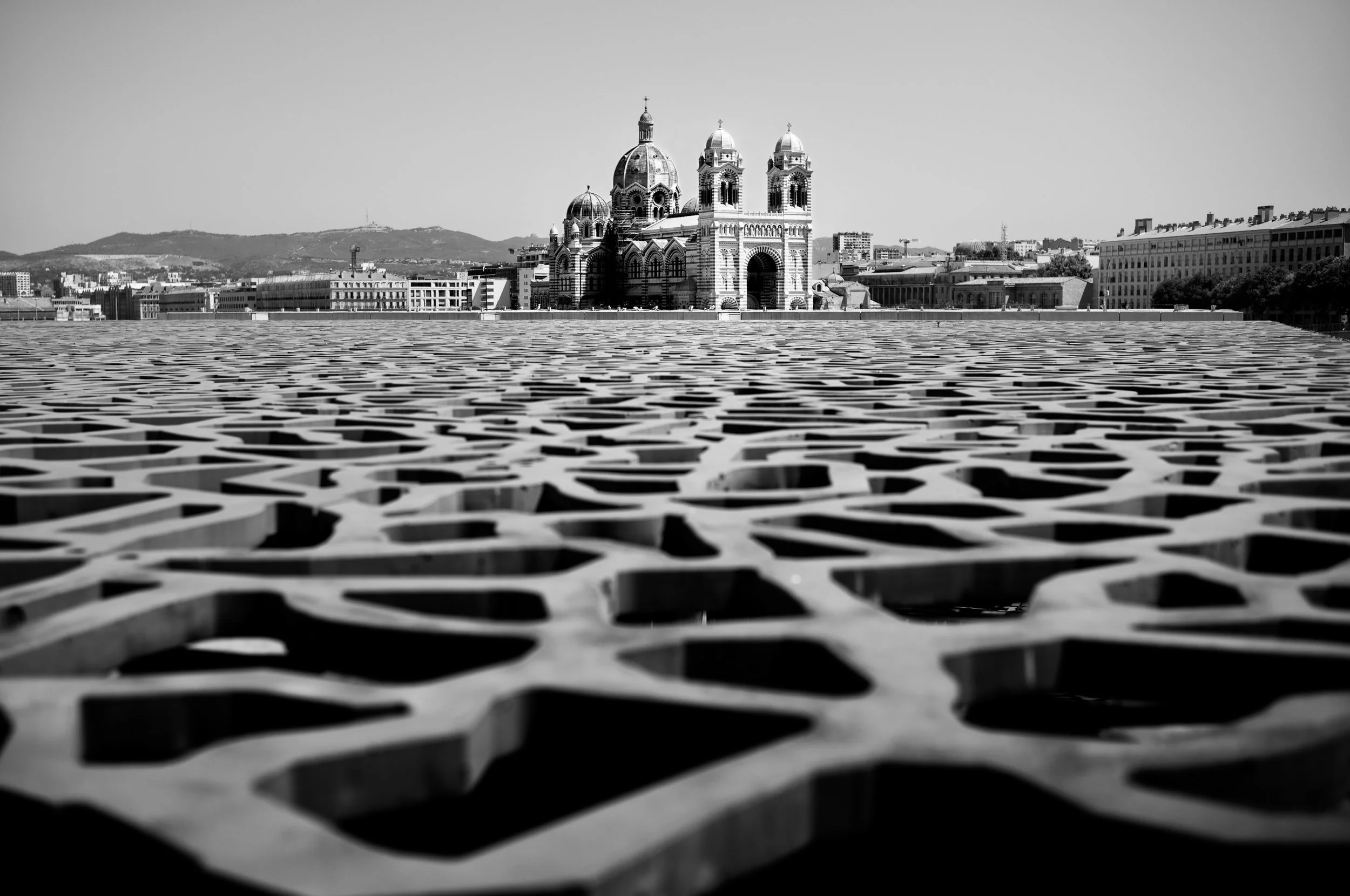 A black and white photograph showing a cityscape with a historic cathedral in the background and an ornate, patterned metal surface in the foreground.