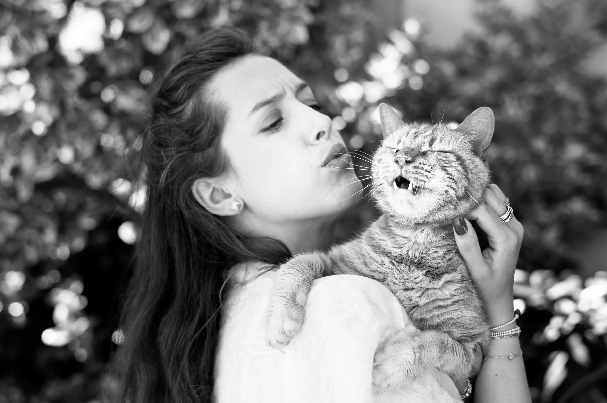 A woman with long dark hair holding a smiling tabby cat with bared teeth, outdoors with blurred foliage in the background.