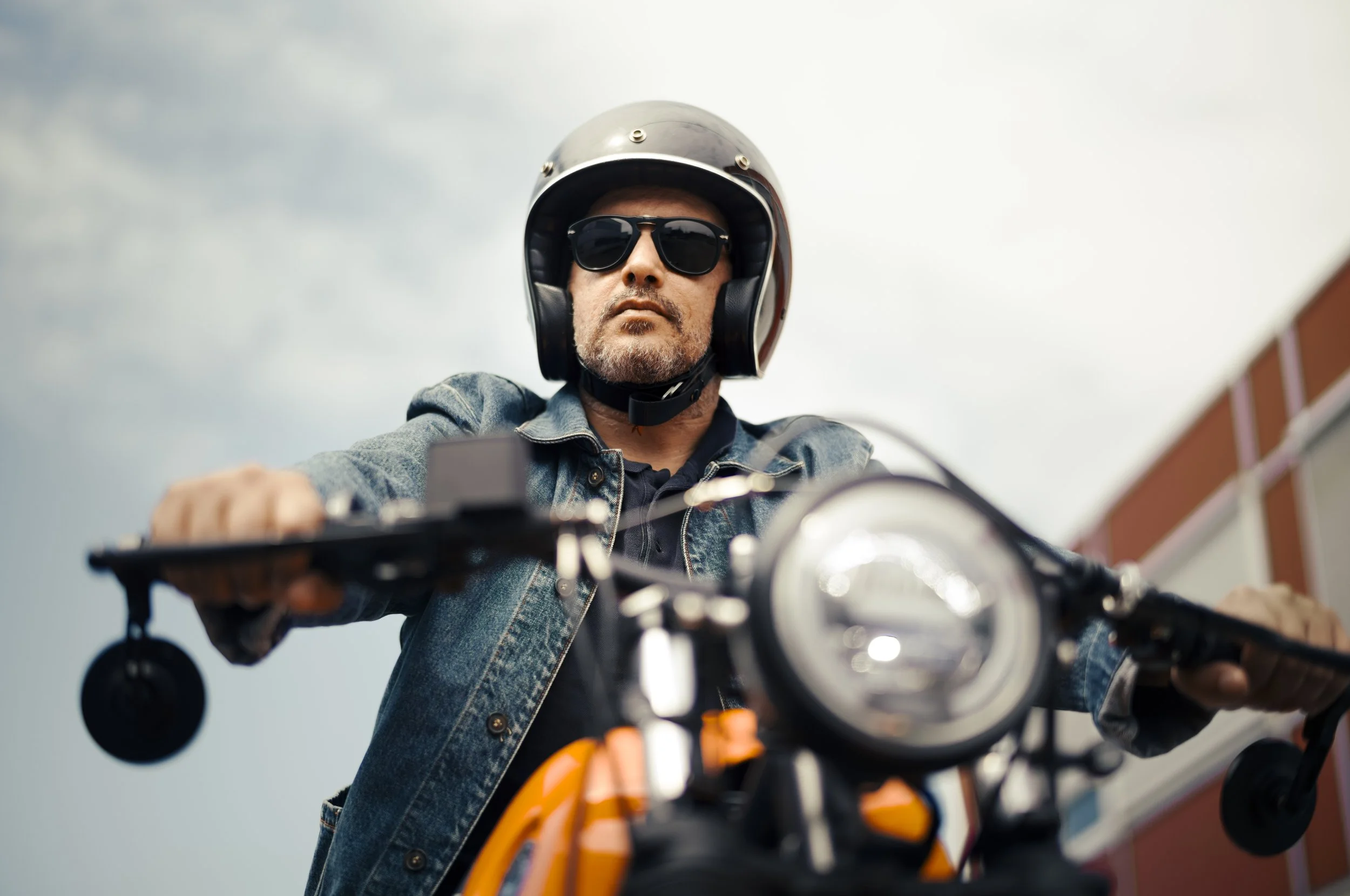 Man wearing sunglasses and a helmet riding a motorcycle, gravelly building and cloudy sky in background