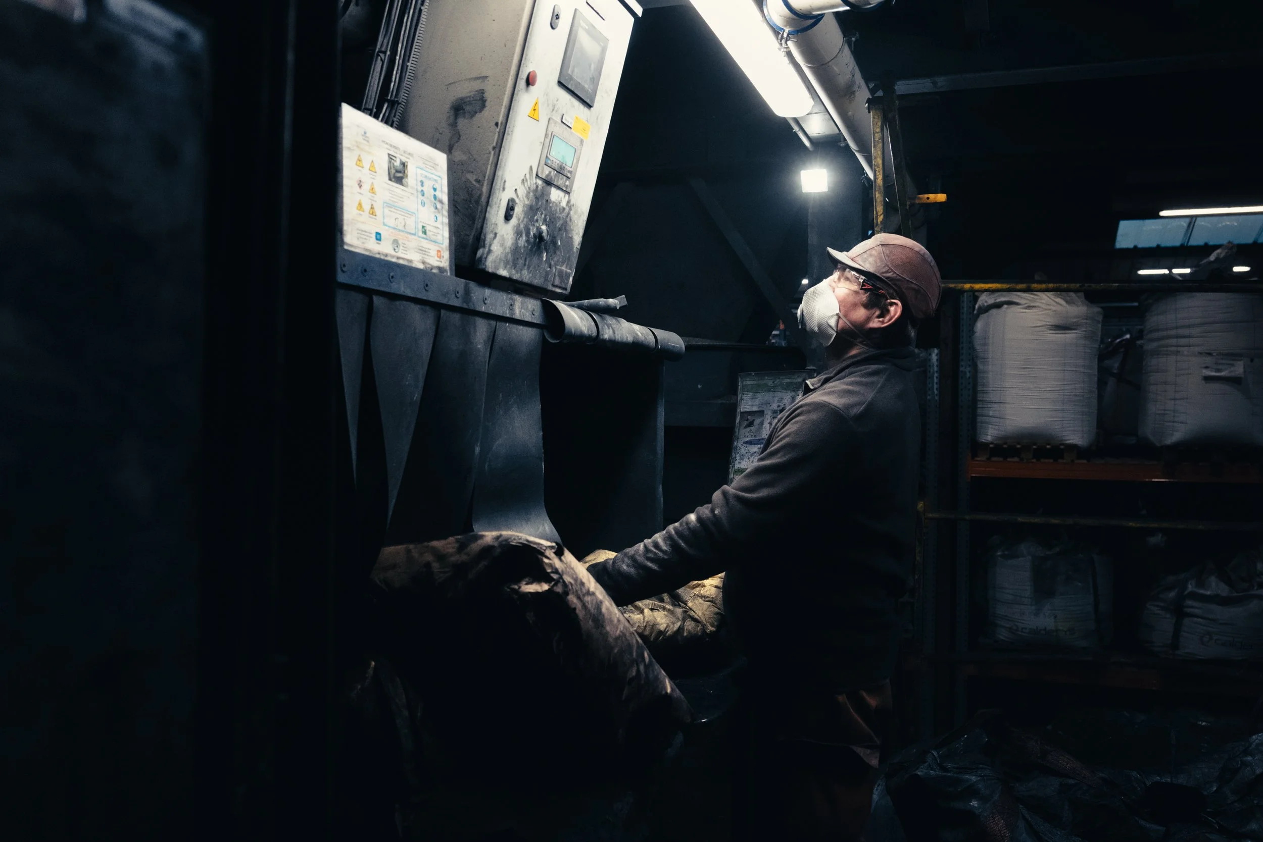 A worker in a dimly lit industrial setting wearing a face mask, glasses, and a cap, operating machinery.