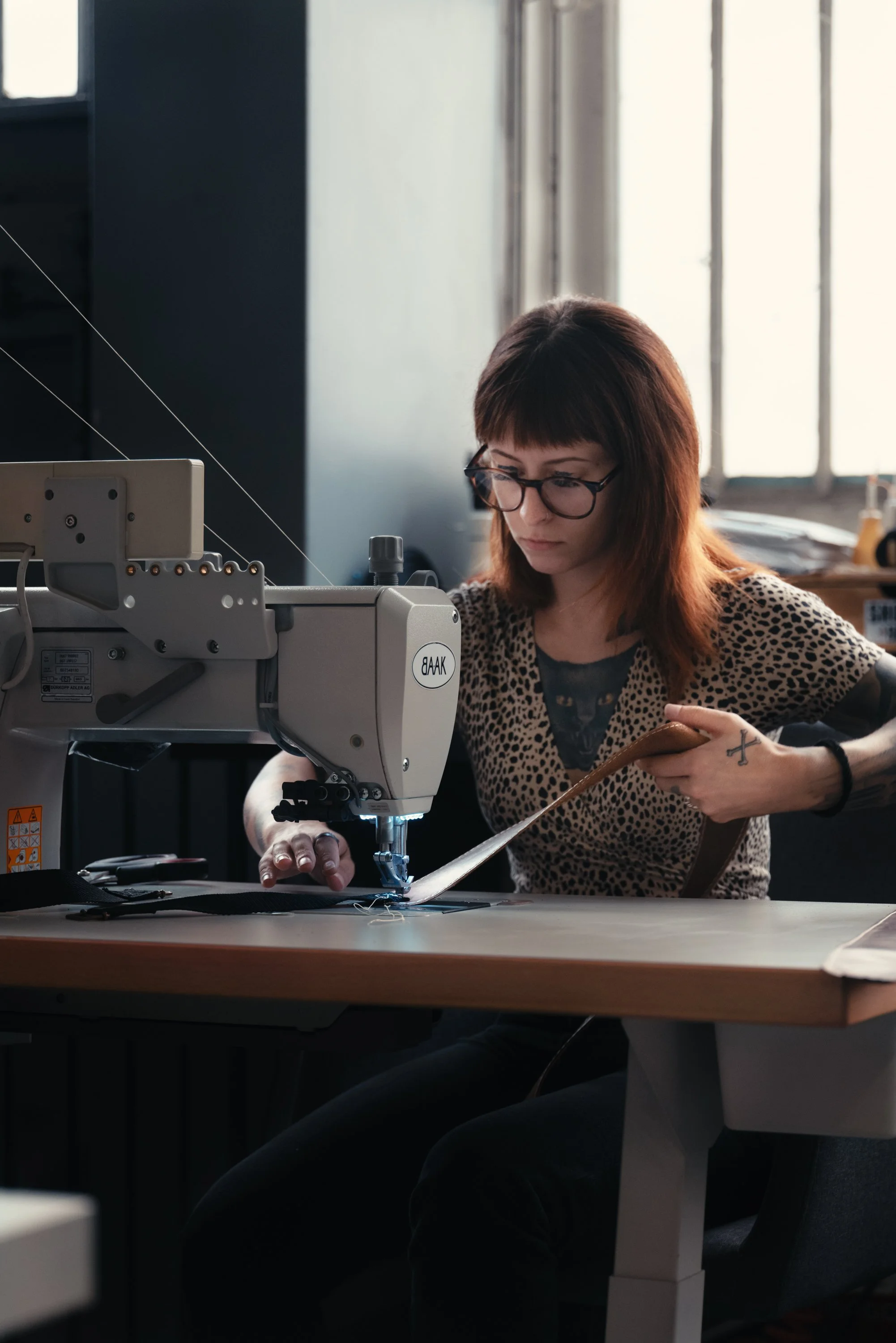 A woman with glasses sewing fabric using a sewing machine in a studio or workshop.