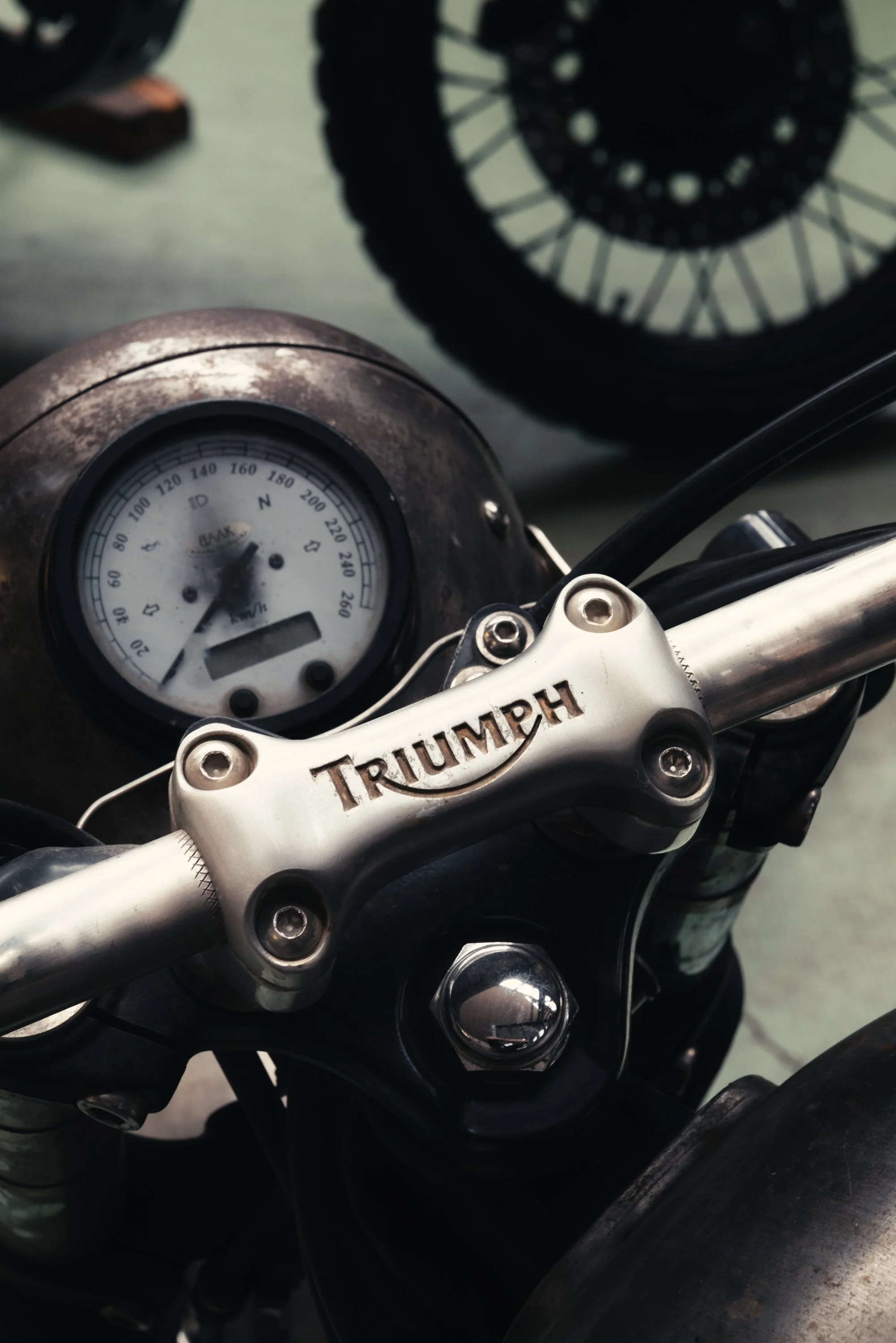Close-up of a Triumph motorcycle handlebar with a silver handlebar clamp and a speedometer in the background, showing a vintage look.