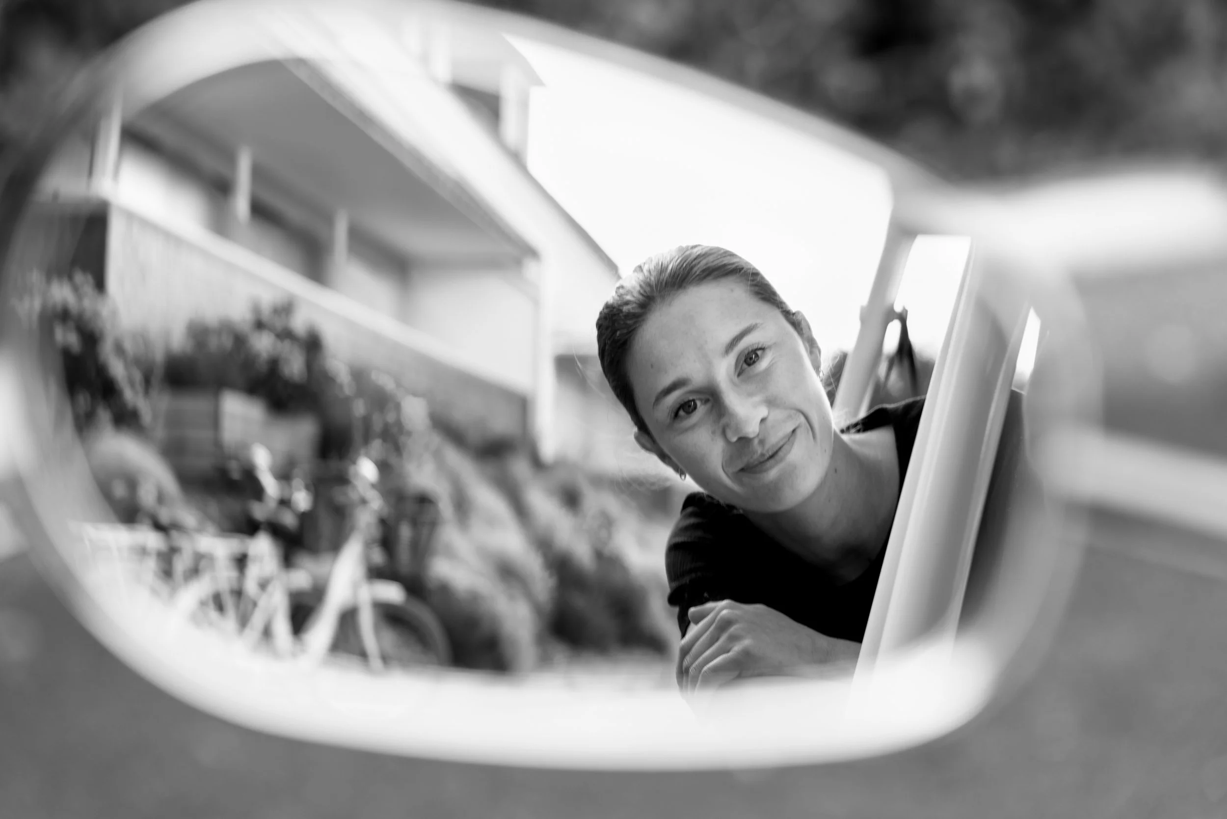 A woman smiling and looking into a mirror with her reflection visible, with outdoor background including a bicycle and plants, in black and white.