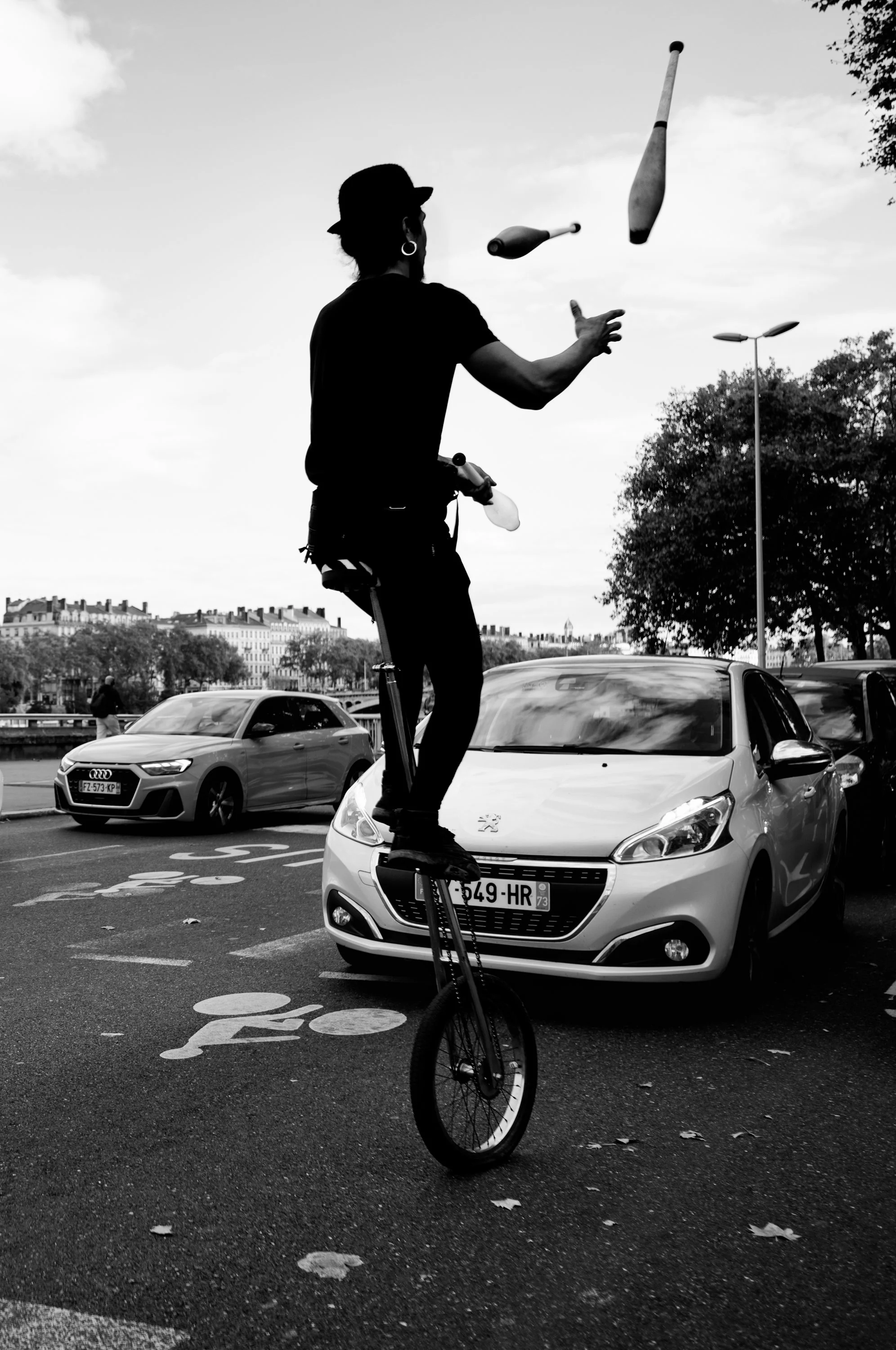 A person performing a unicycle act by juggling three clubs on a street in front of parked cars with a cityscape and trees in the background.