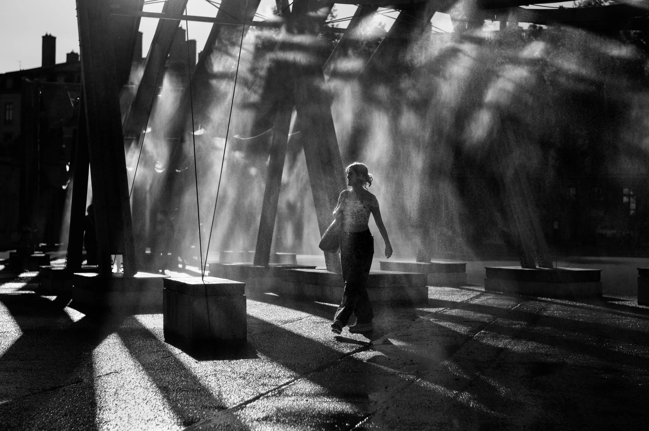 A person walking through a playground with swings, shadows cast by the sunlight, and dust or mist in the air in black and white.