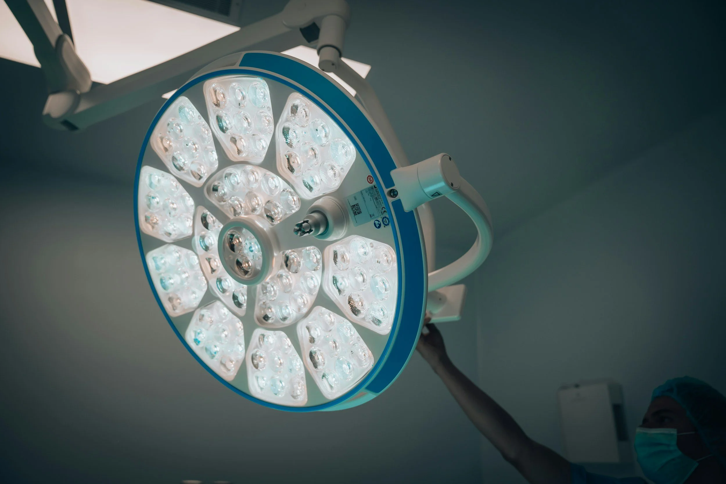 A large circular surgical LED light in an operating room, with a healthcare worker wearing a mask and hair covering adjusting it.