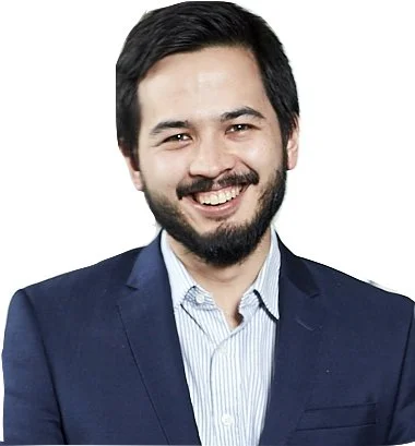 A man with dark hair and a beard, wearing a blue blazer and a light blue collared shirt, smiling against a white background.