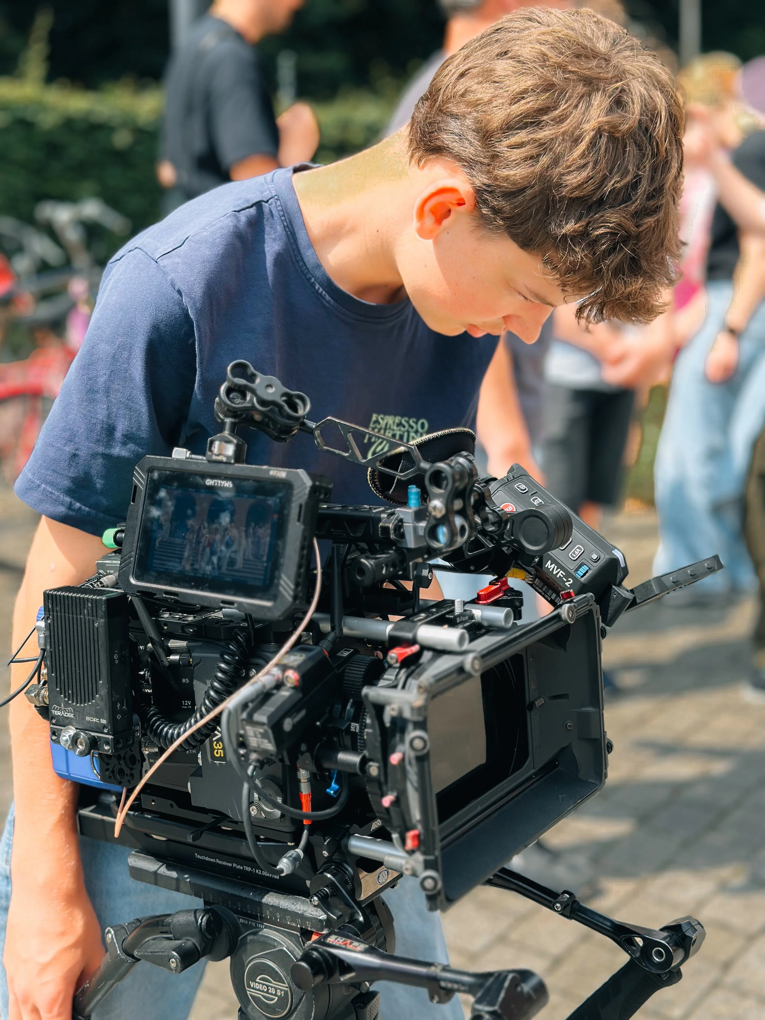 A person is operating a professional film camera outdoors, with other people blurred in the background.