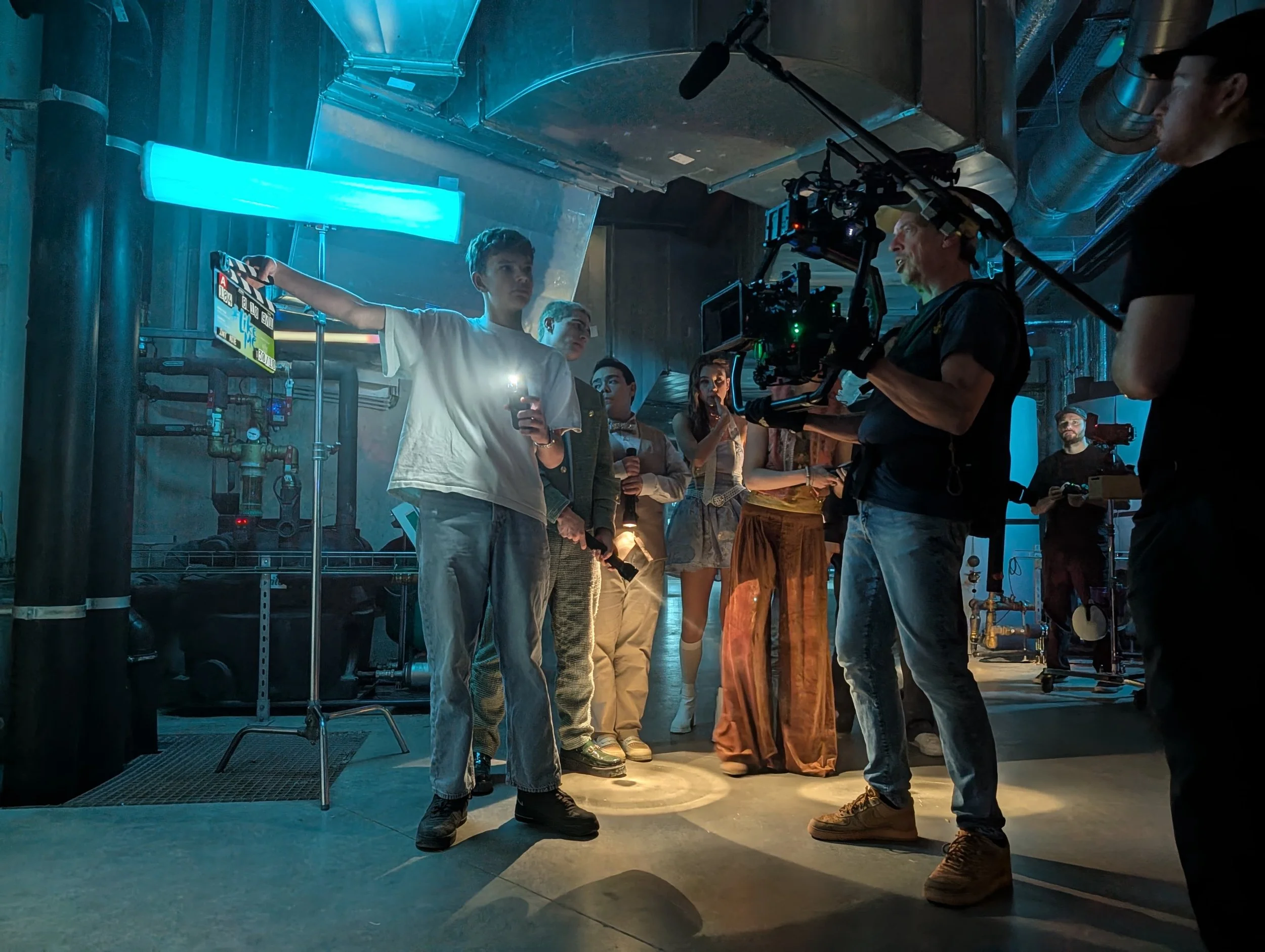 Behind the scenes of a film or TV show set, a young man in a white t-shirt holds a clapperboard next to a woman with long hair, and a camera operator films them. Several crew members are present, and the environment is industrial with pipes and blue lighting.