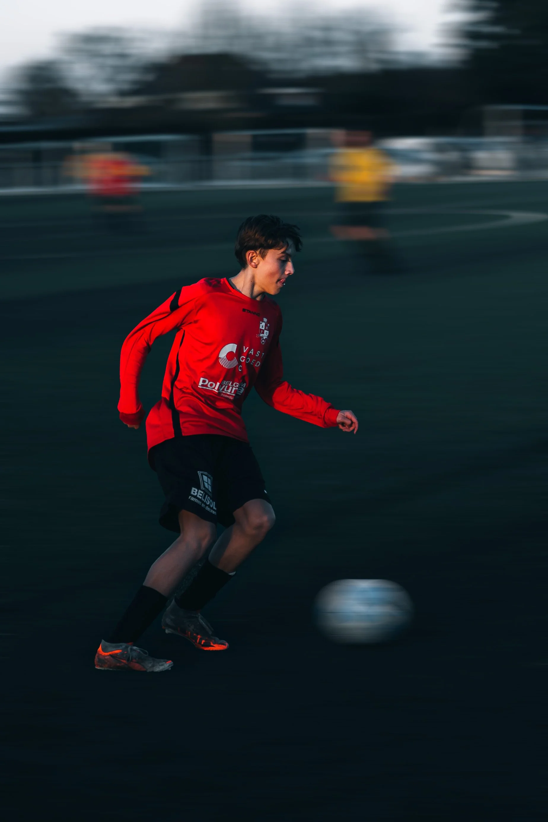A young male soccer player in a red jersey running on a dark field during a match. The background shows blurred figures of other players and spectators.