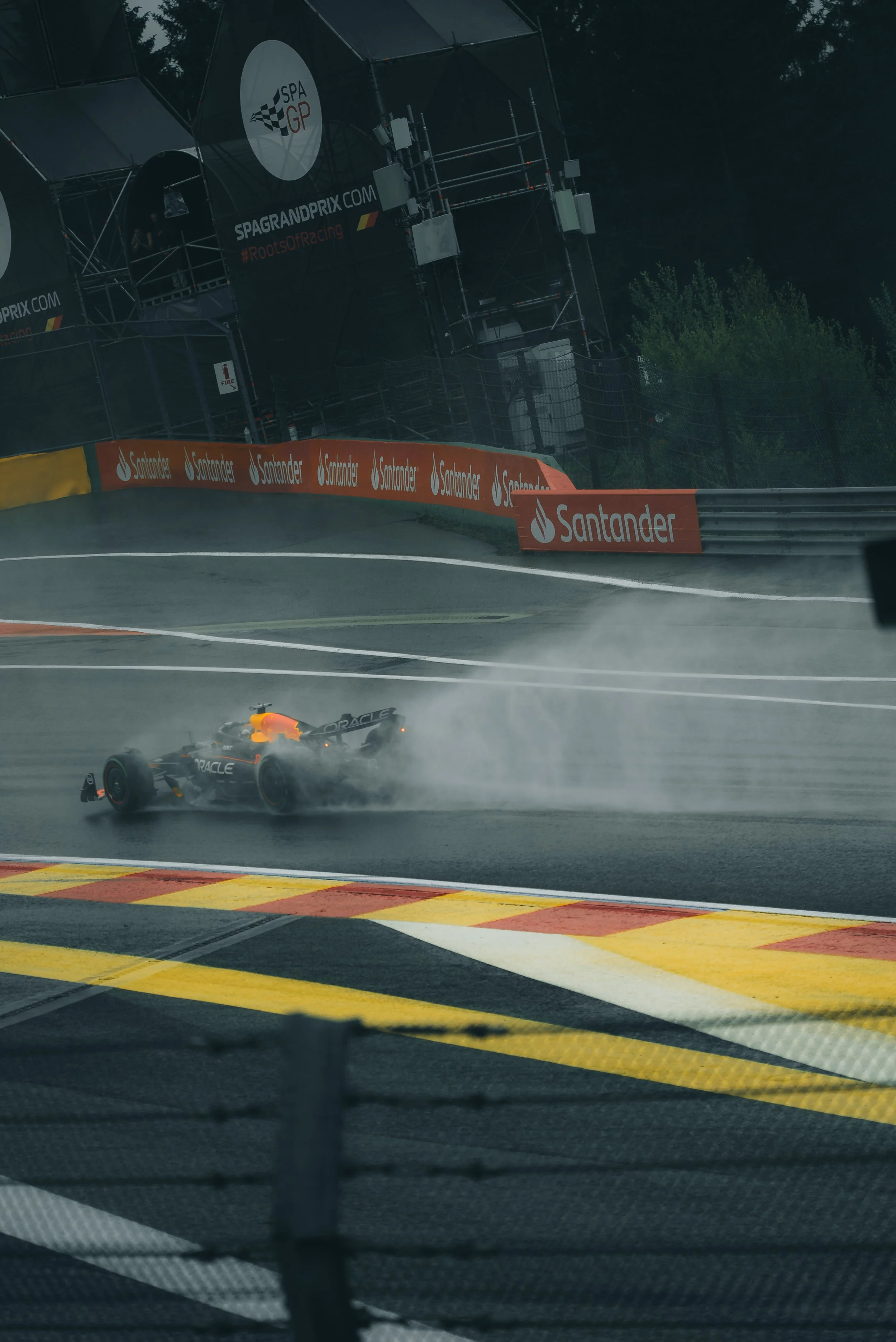 A formula race car on a wet race track with spray behind it, approaching a turn. The scene is viewed from inside a vehicle, with parts of the dashboard and track markings visible. There are advertising banners along the track, including Santander.