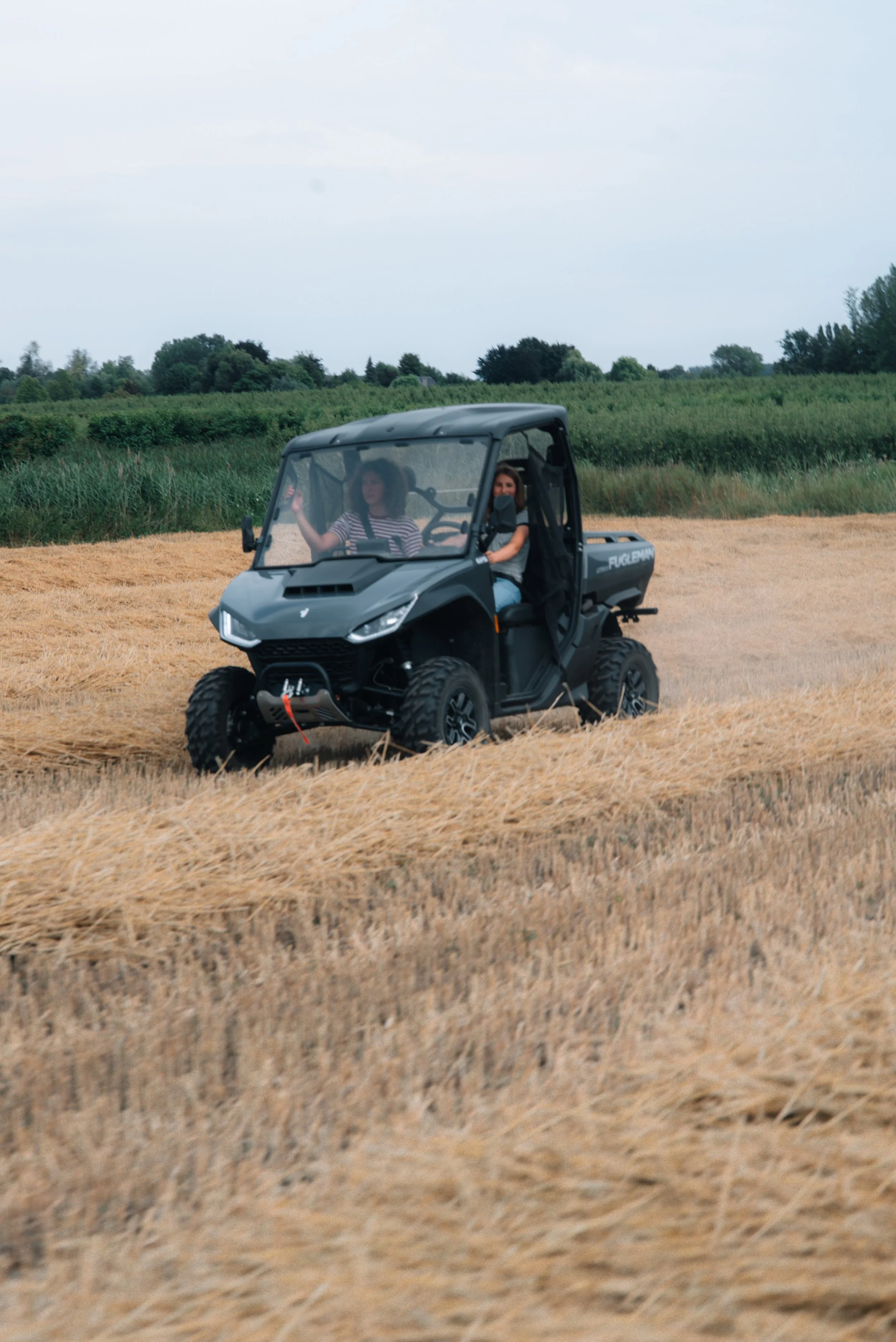 Two women riding in a black utility vehicle through a field of dry grass with green trees and a cloudy sky in the background.
