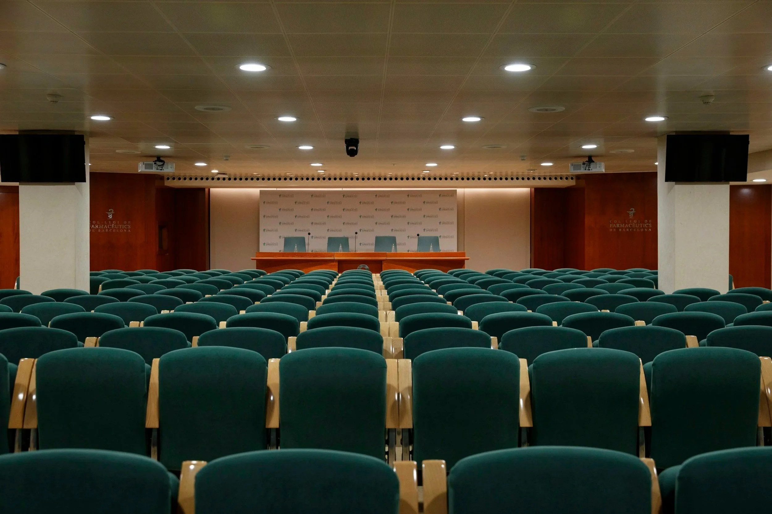 An empty lecture hall with rows of green chairs facing a stage with a backdrop displaying a logo. The hall has a wooden ceiling with multiple circular overhead lights and two wall-mounted screens.