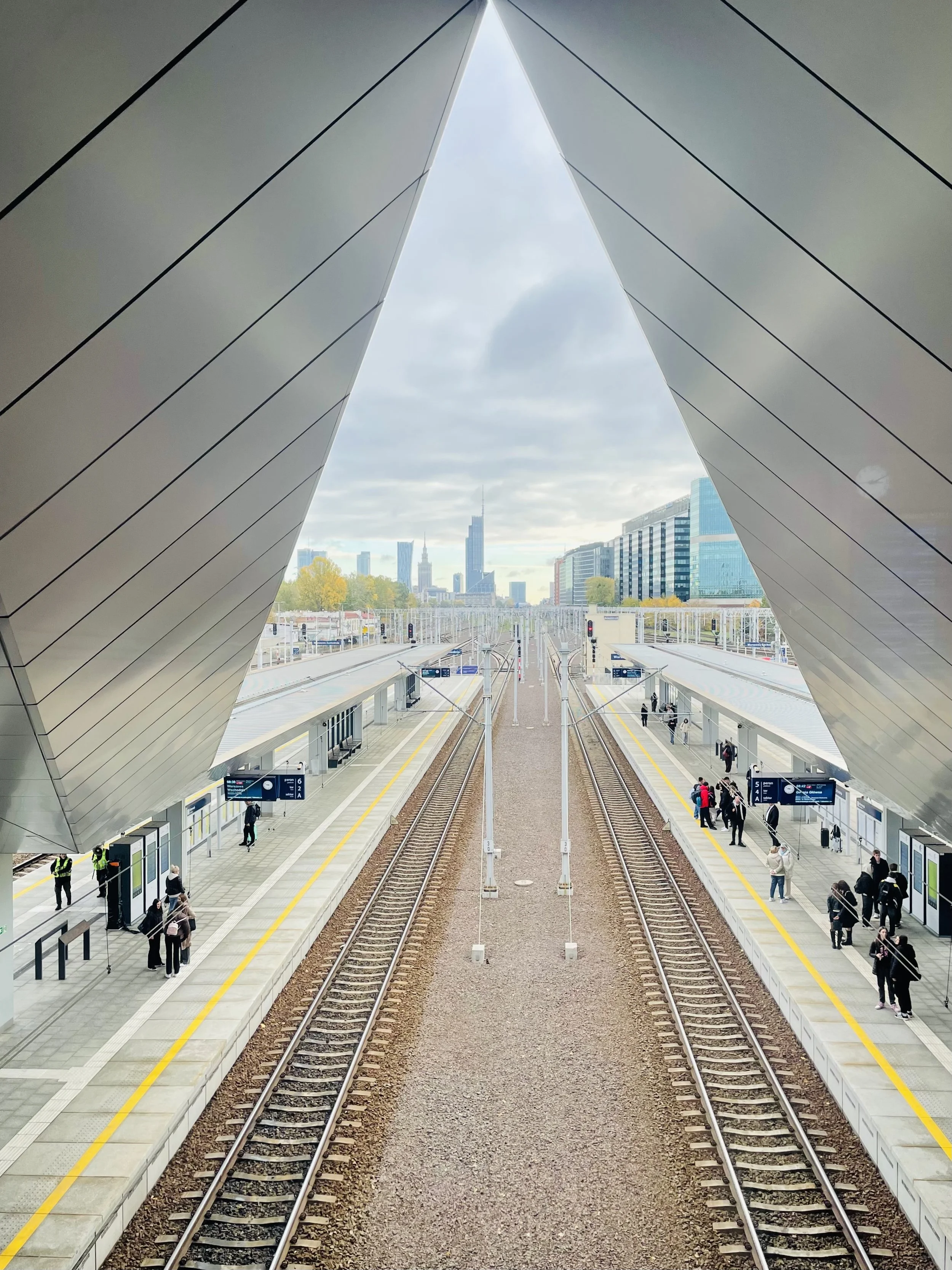 View of a modern train station with multiple train tracks, platforms, and passengers waiting. A city skyline with tall buildings is visible in the background through an architectural structure with a large triangular opening.