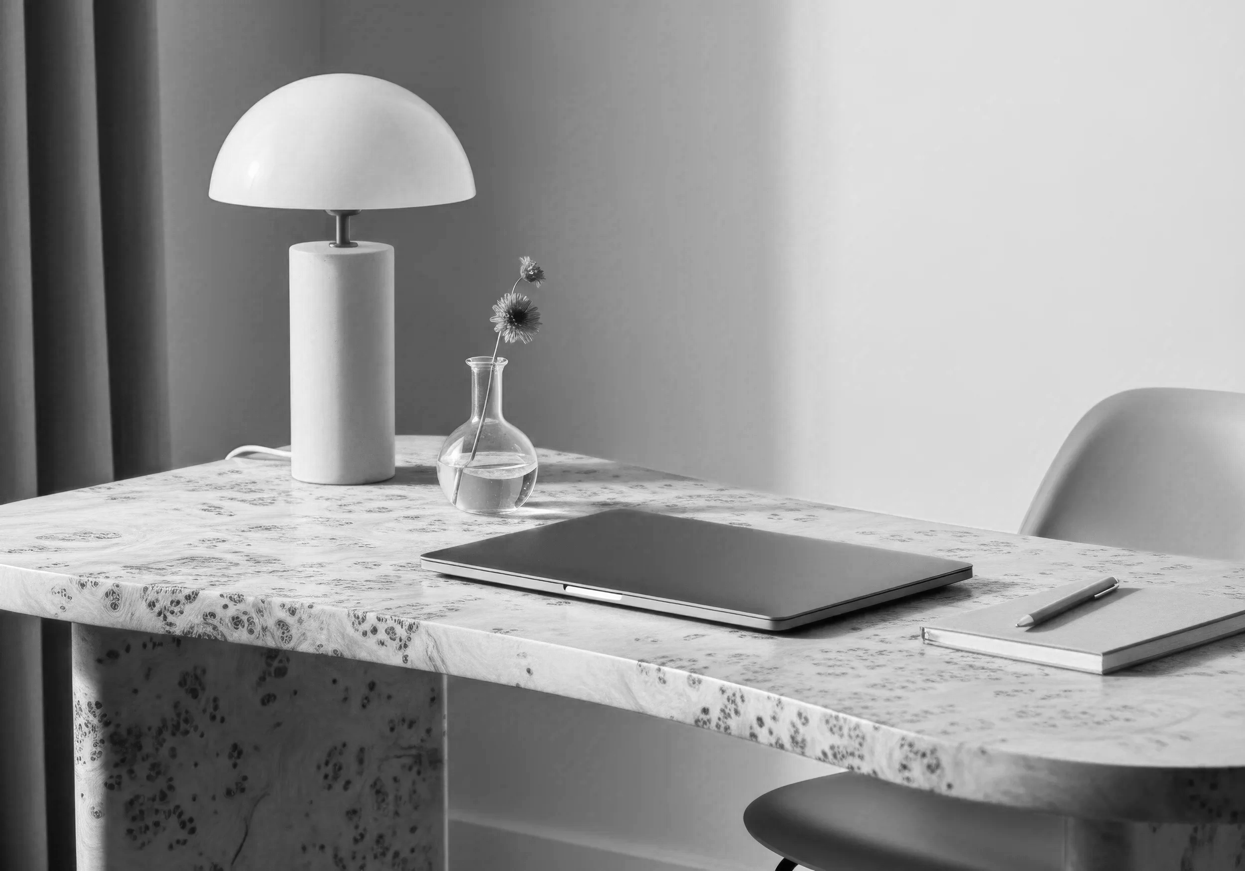A minimalist workspace with a marble table, a closed laptop, a pen, a small vase with dried flowers, a table lamp, and a chair against a wall with curtains.