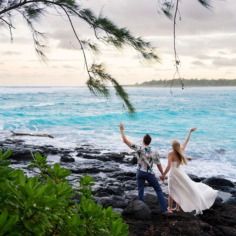 A couple holding hands on a rocky shoreline, facing the ocean, with the woman in a white dress and the man in casual attire, under a cloudy sky.