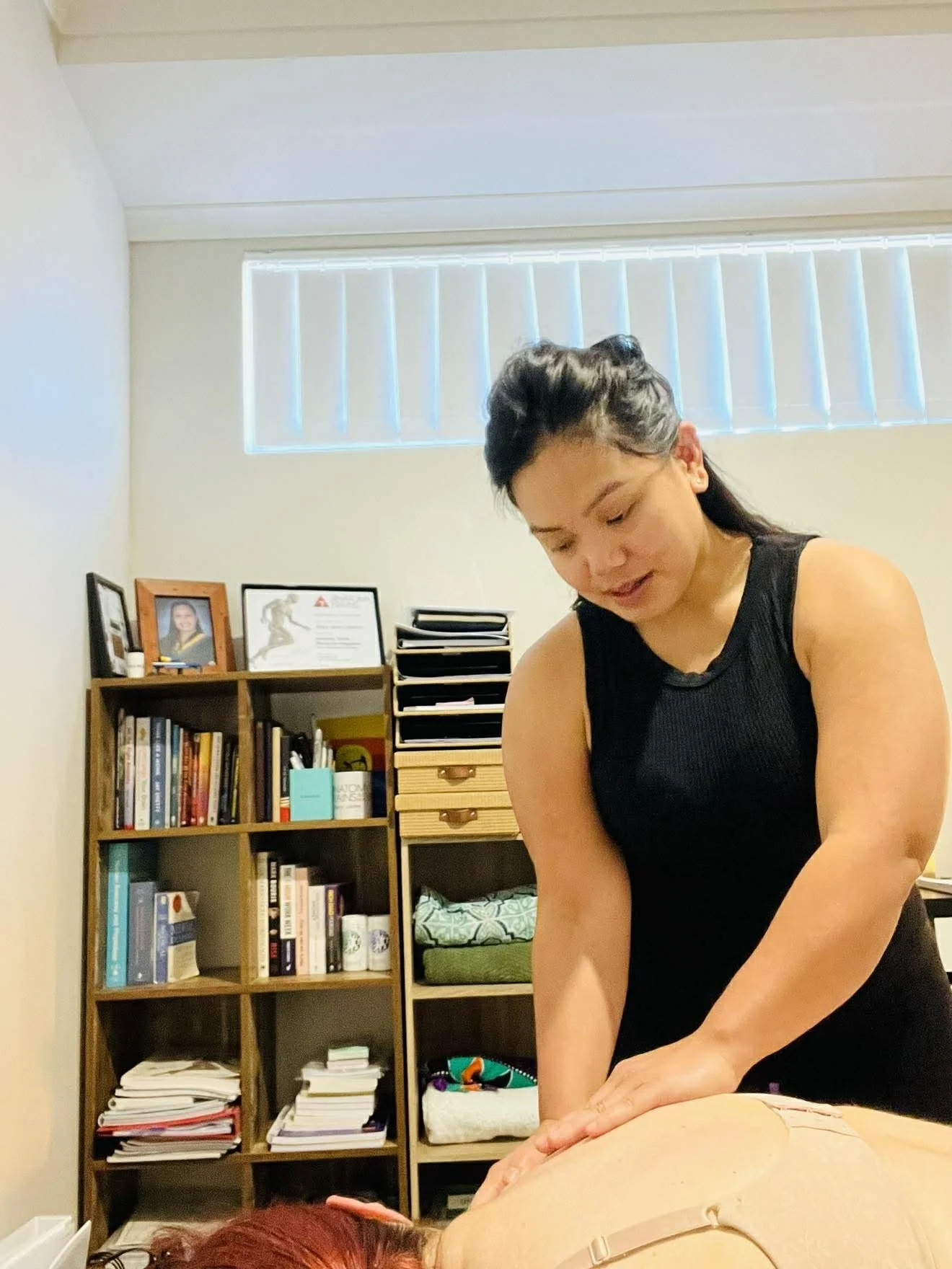 A woman giving a massage to a person lying face down on a table in a room with a bookshelf in the background.