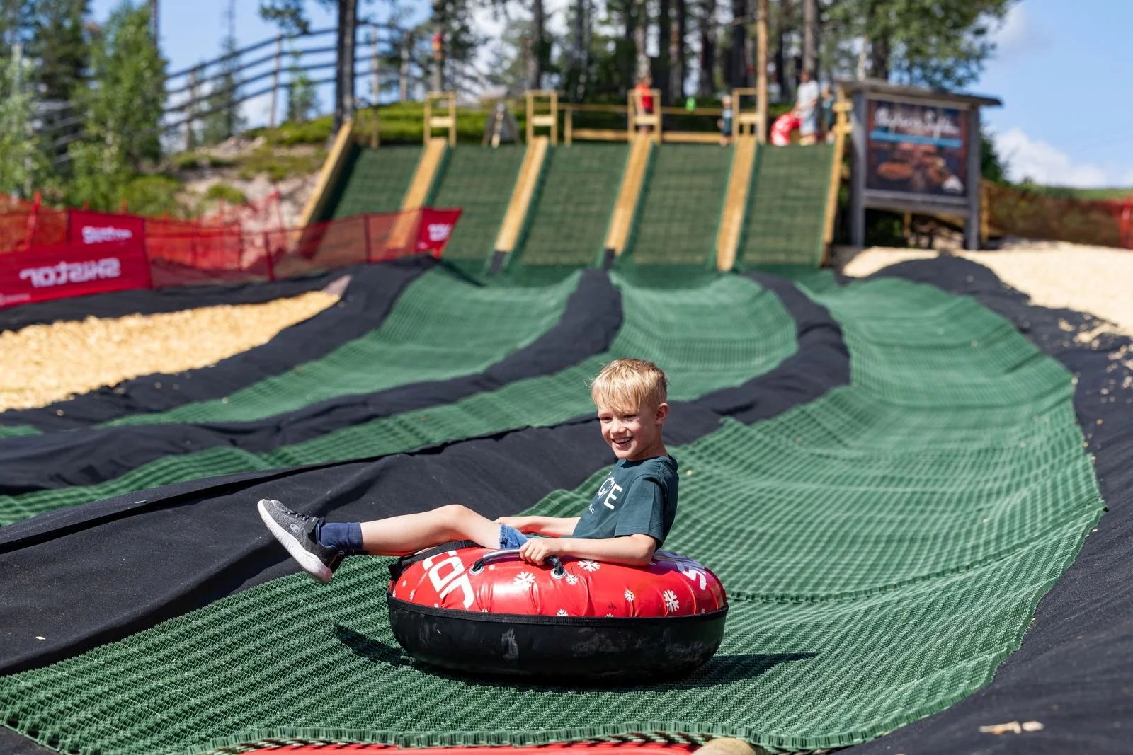 Uppbyggnadsarbete vid en skidbacke med gröna matta och röda avspärrningsstängsel, med en restaurang i träbyggnad och omgivande träd och hus i bakgrunden.