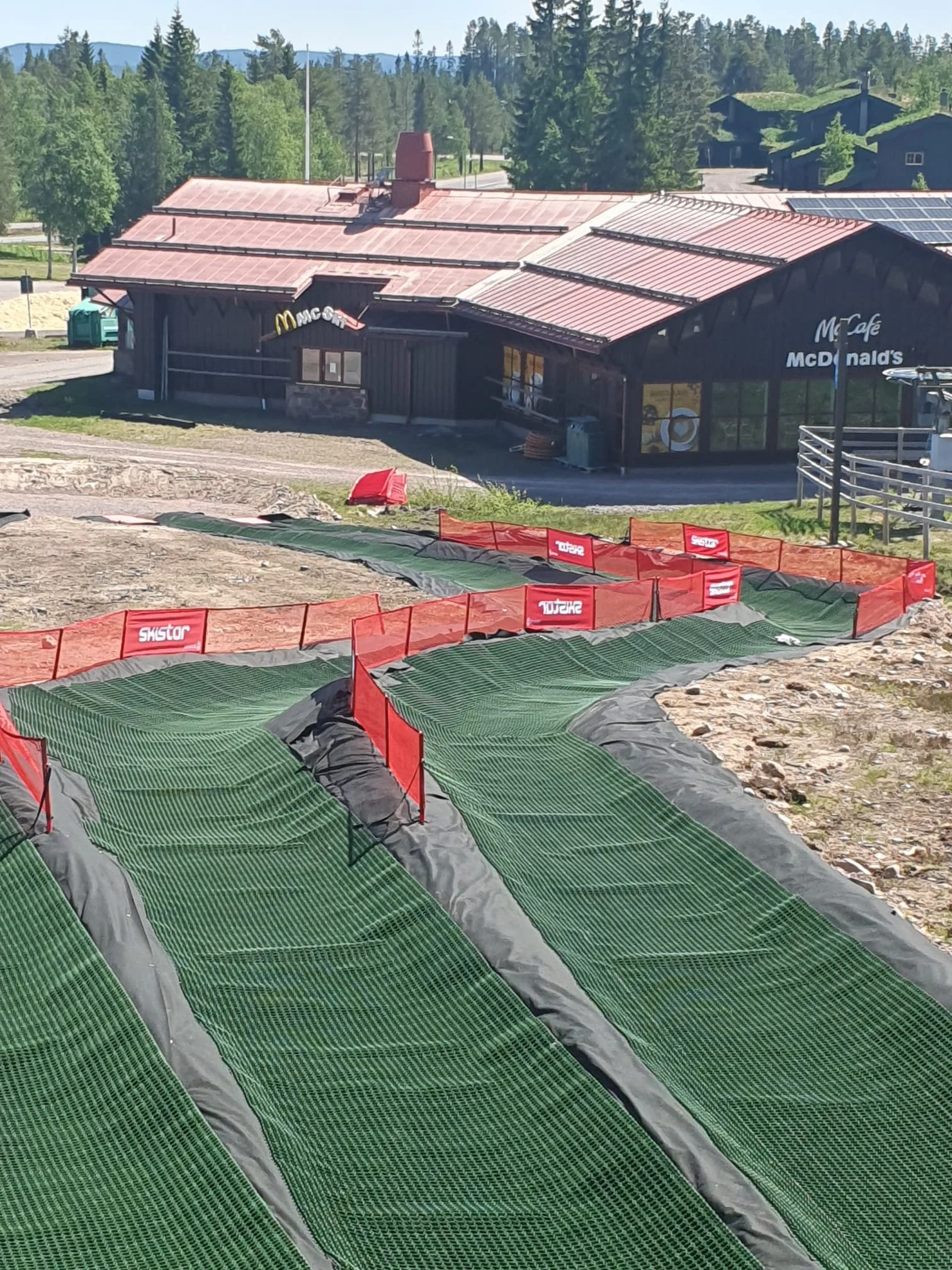 Uppbyggnadsarbete vid en skidbacke med gröna matta och röda avspärrningsstängsel, med en restaurang i träbyggnad och omgivande träd och hus i bakgrunden.