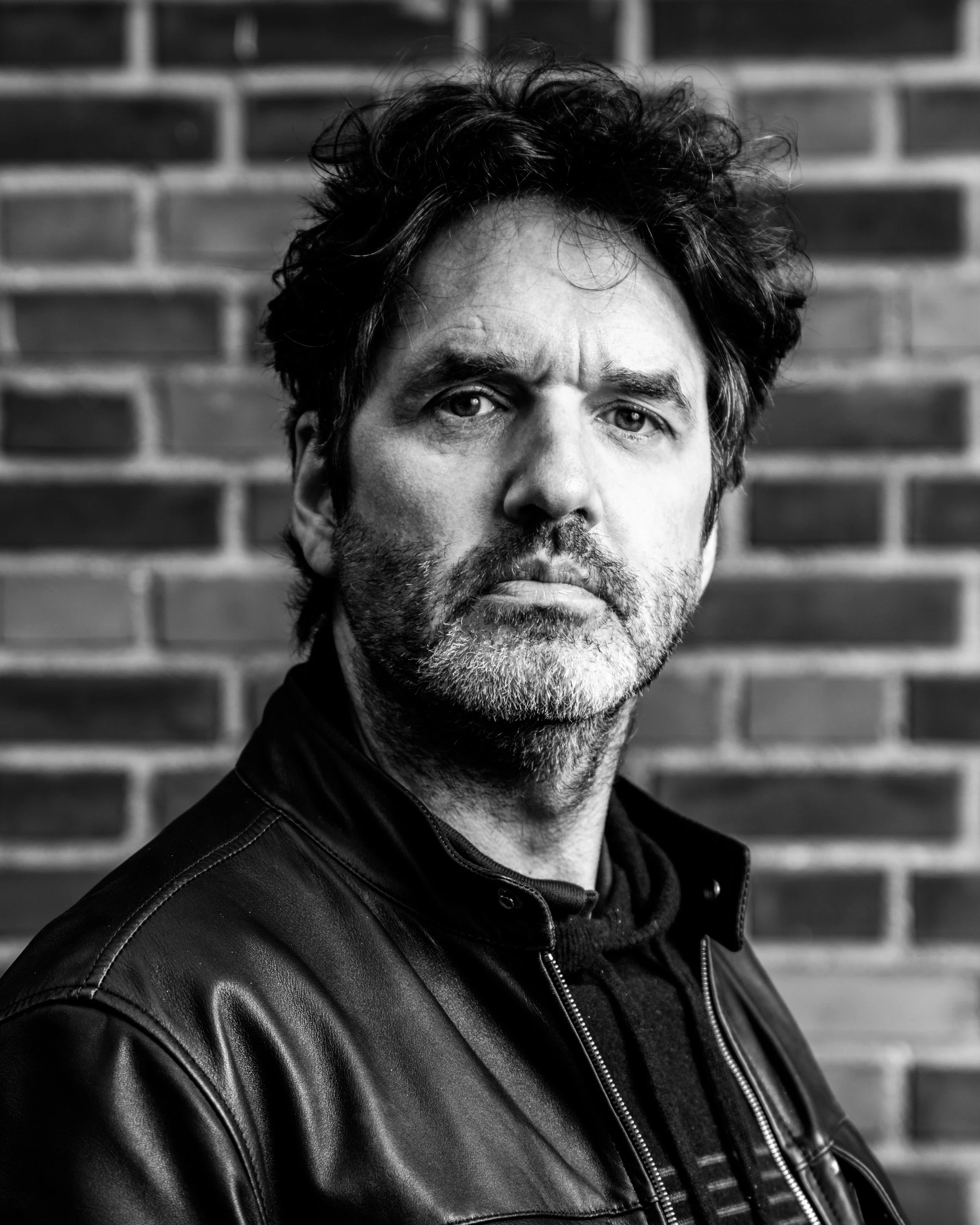 Black and white portrait of a middle-aged man with dark wavy hair and stubble, wearing a leather jacket, standing against a brick wall.