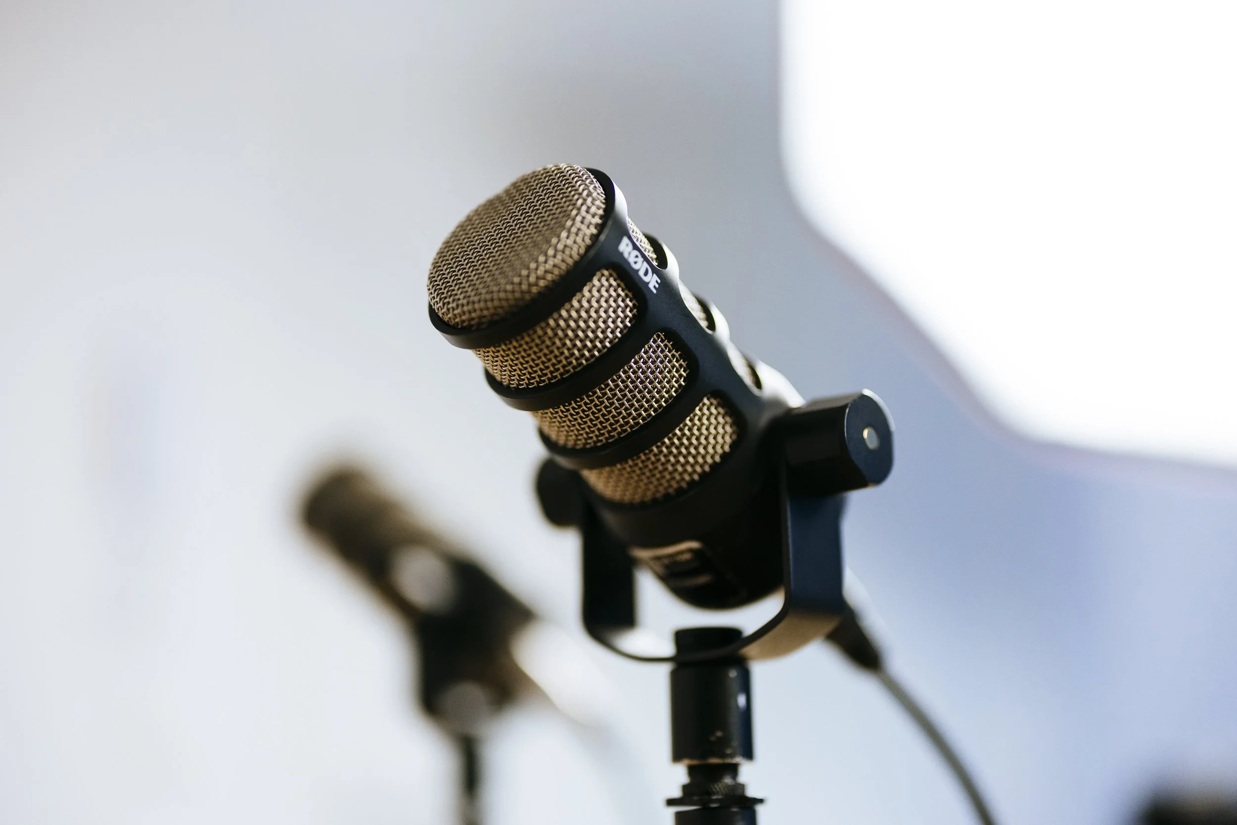 A close-up of a professional podcast microphone on a stand, with another microphone blurred in the background.