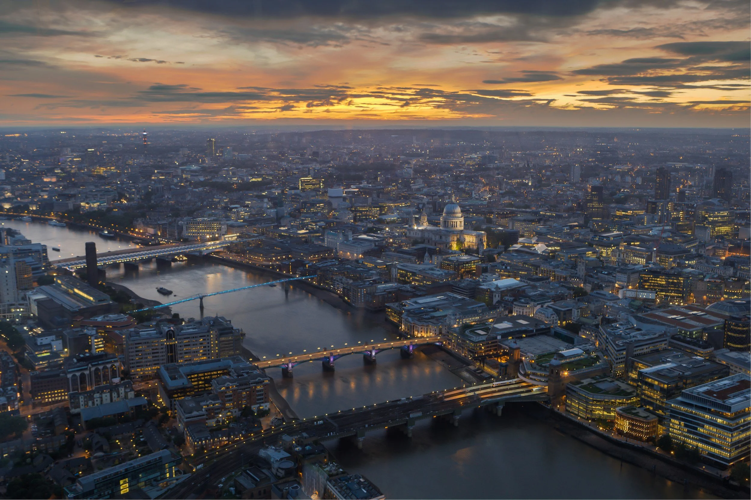 Aerial view of London at sunset showing illuminated bridges over the River Thames, historic and modern buildings, and a dramatic sky with clouds.