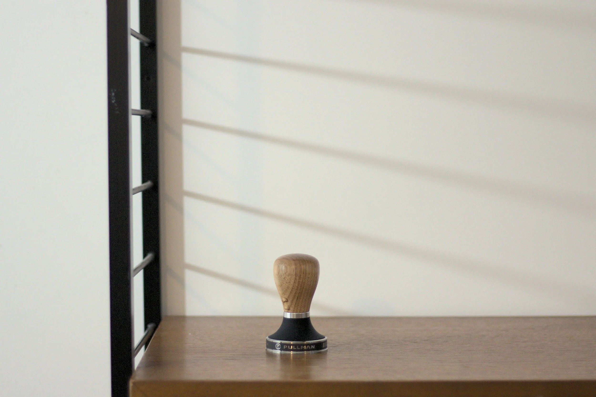 A wooden-handled coffee tamper on a wooden shelf with a white wall and shadows in the background.