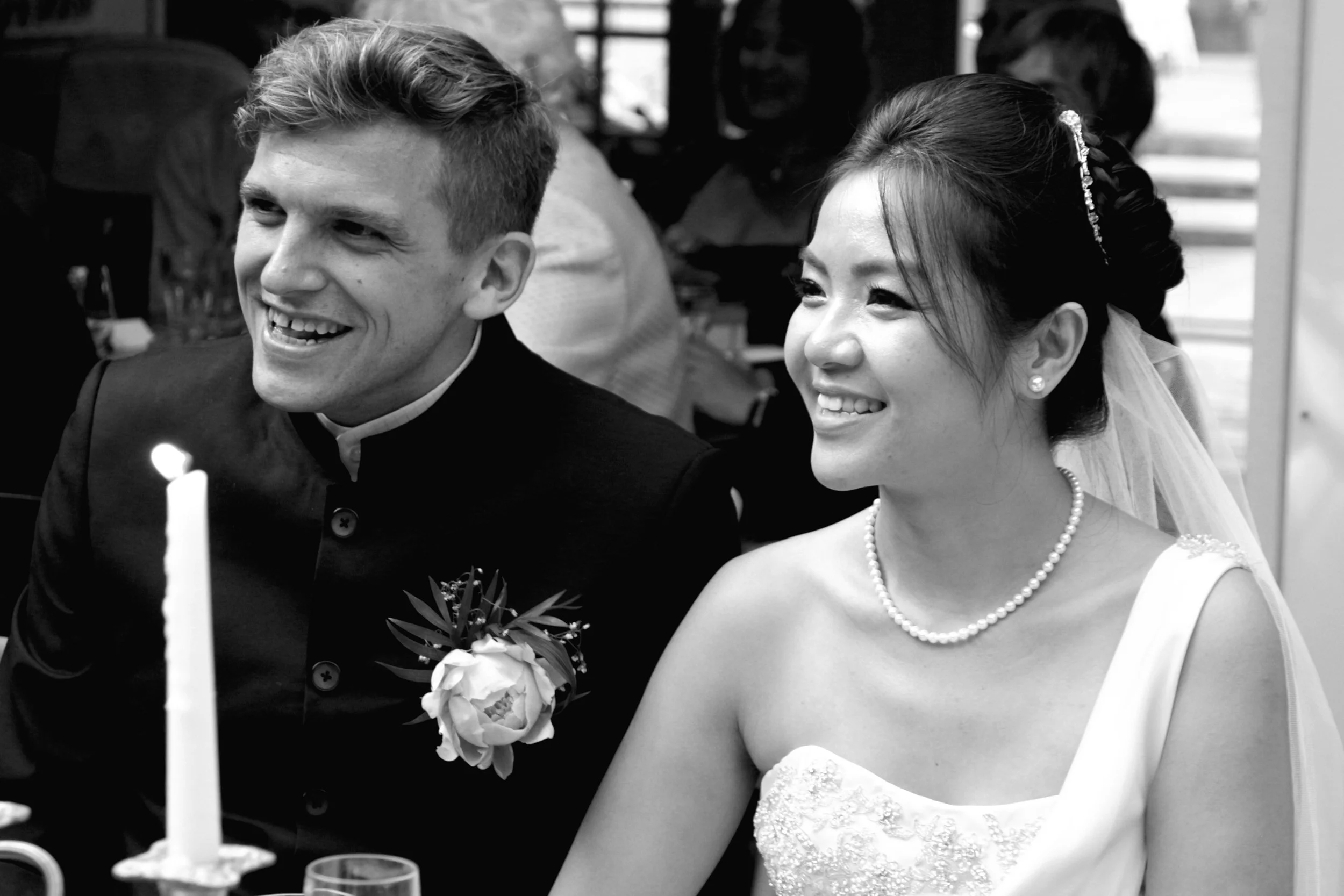 A smiling bride and groom at a wedding reception, sitting at a table with a candle in the foreground.