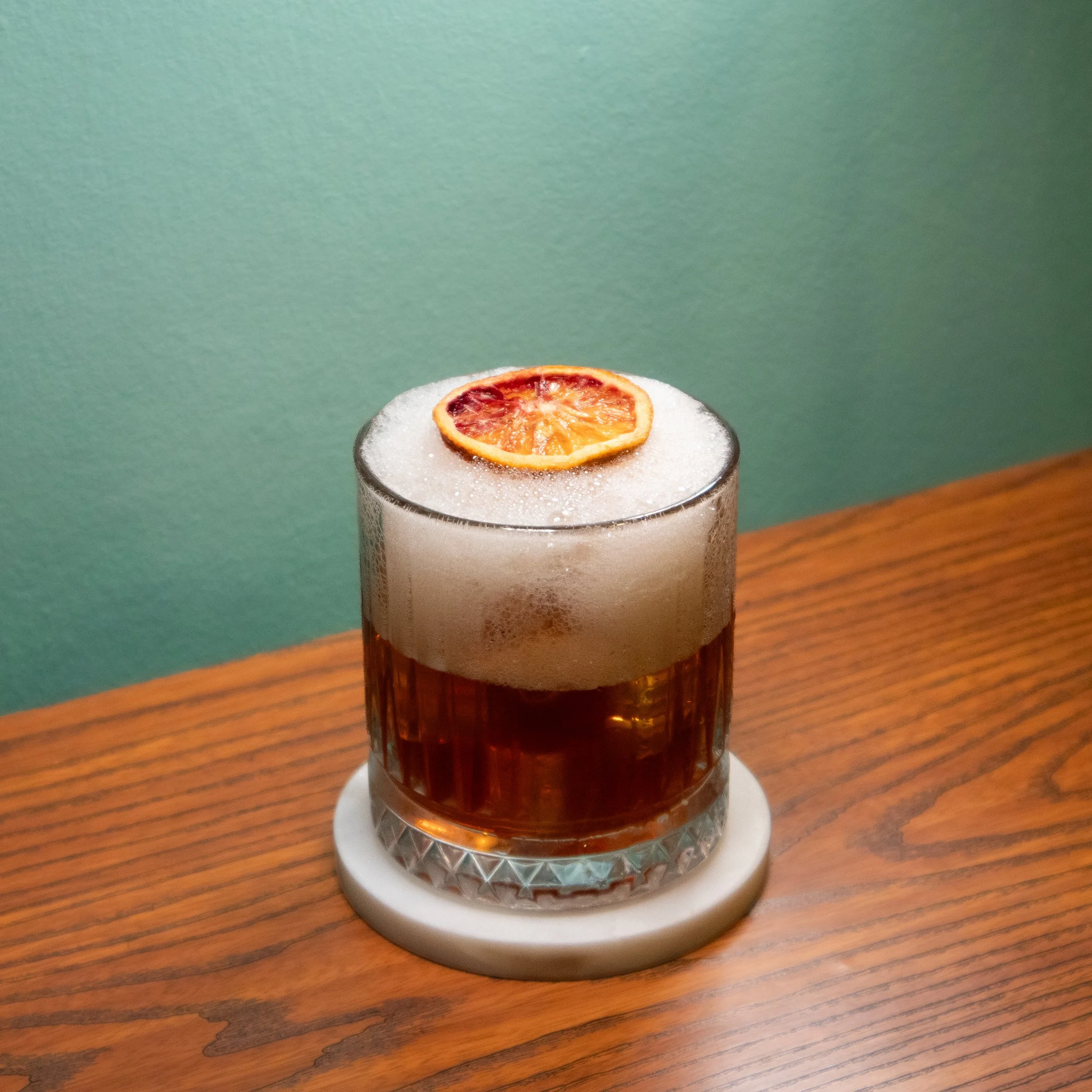 A glass of amber-colored beverage topped with foam and a slice of dried orange, placed on a white decorative coaster on a wooden surface with a green wall in the background.