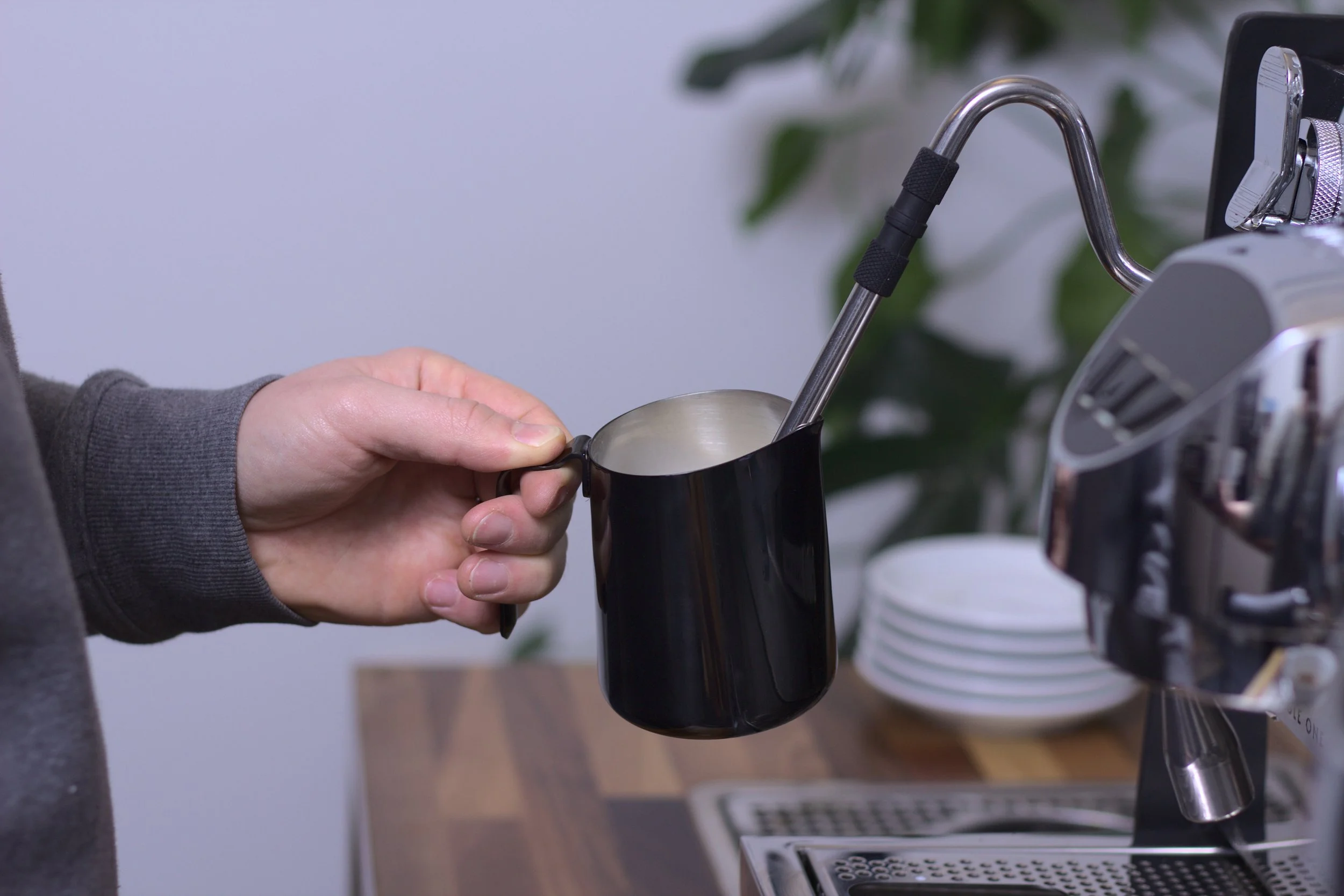 A person holding a black milk pitcher under a coffee machine's steam wand.