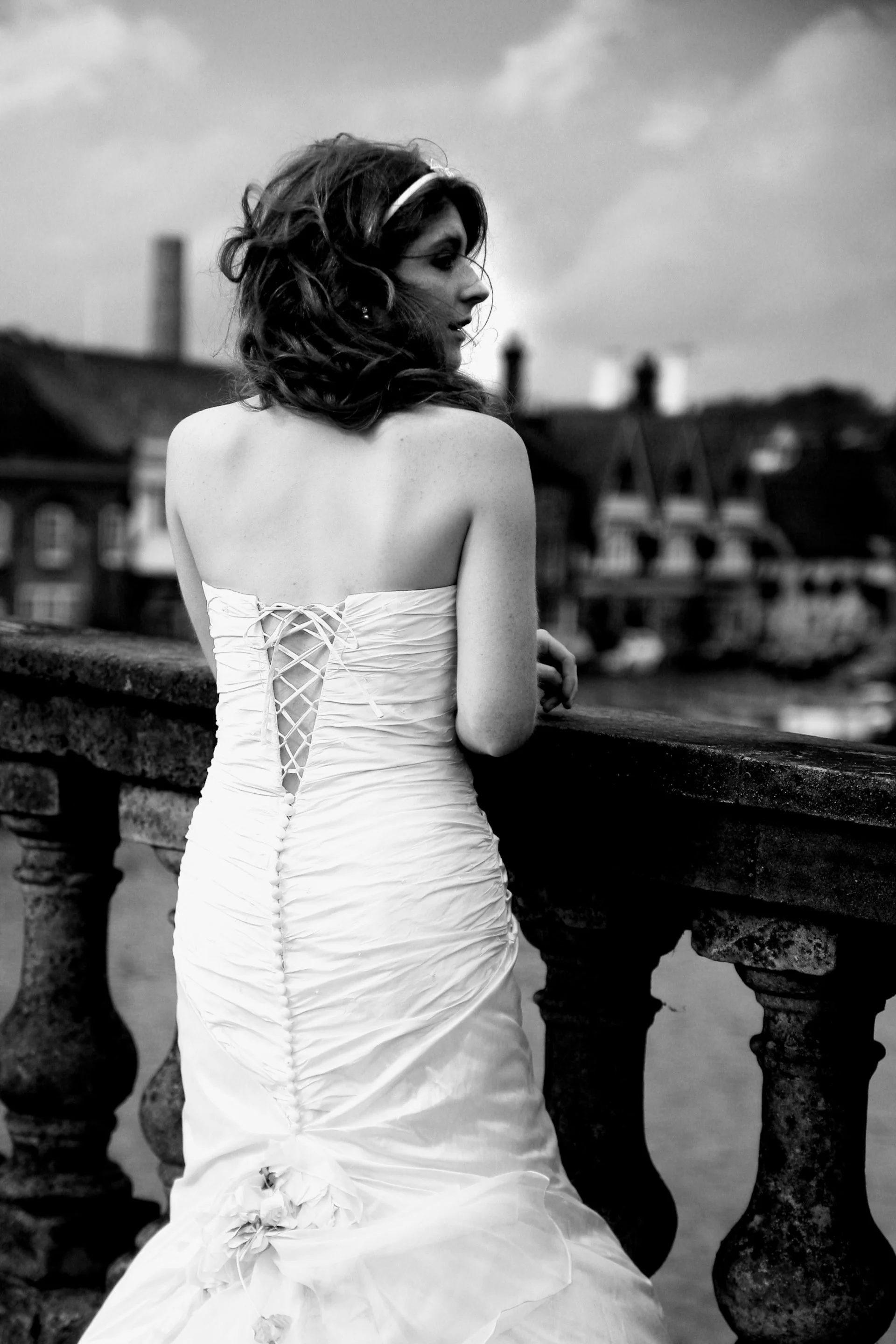 A woman with wavy hair, wearing a strapless wedding dress with lace-up back details, standing by a stone railing with buildings and a cloudy sky in the background.