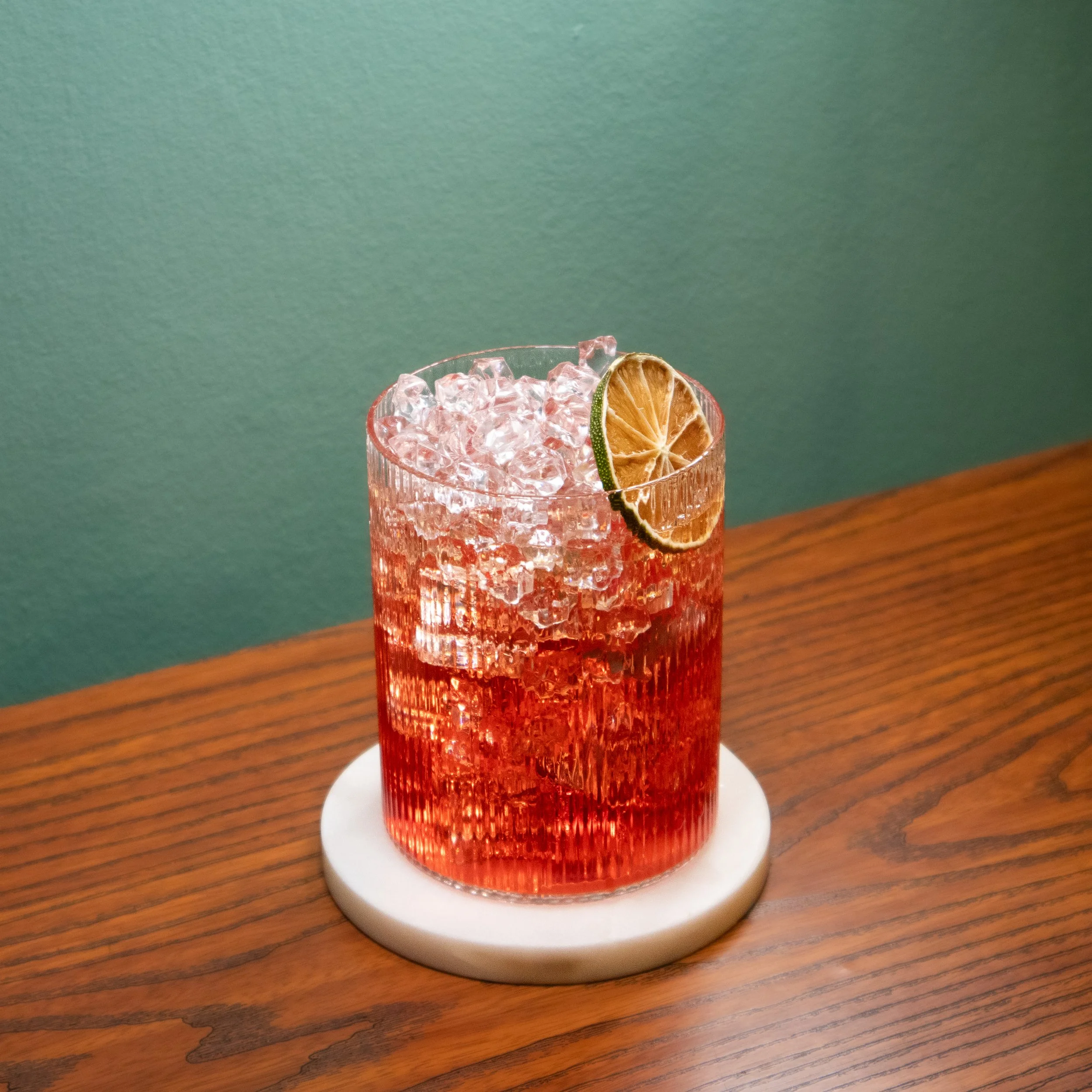 A glass filled with crushed ice and a red liquid, garnished with a dried lime slice, placed on a white decorative coaster on a wooden surface with a green wall in the background.