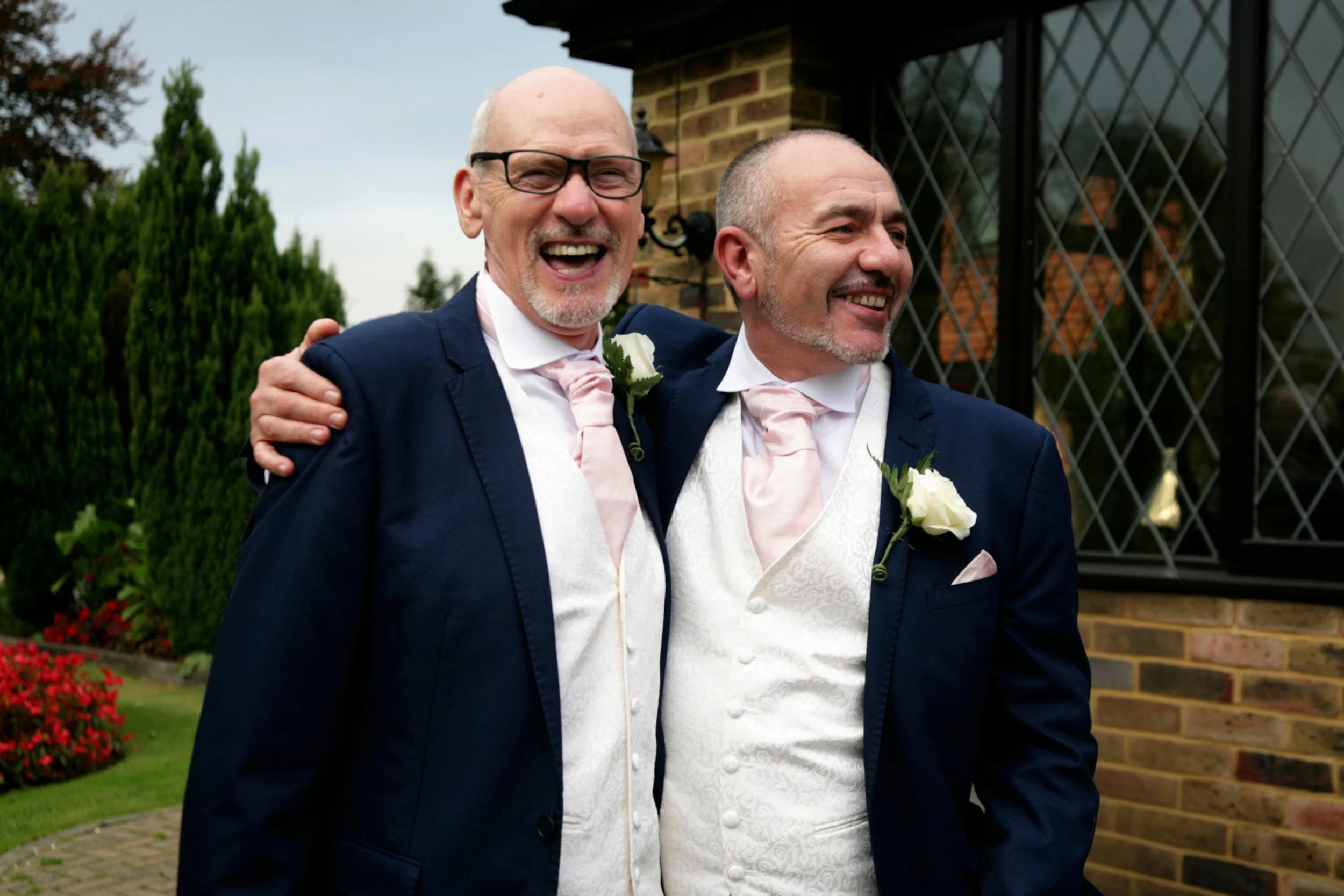 Two men, dressed in tuxedos with pink ties and white shirts, standing outdoors in front of a brick building, smiling and celebrating a wedding.