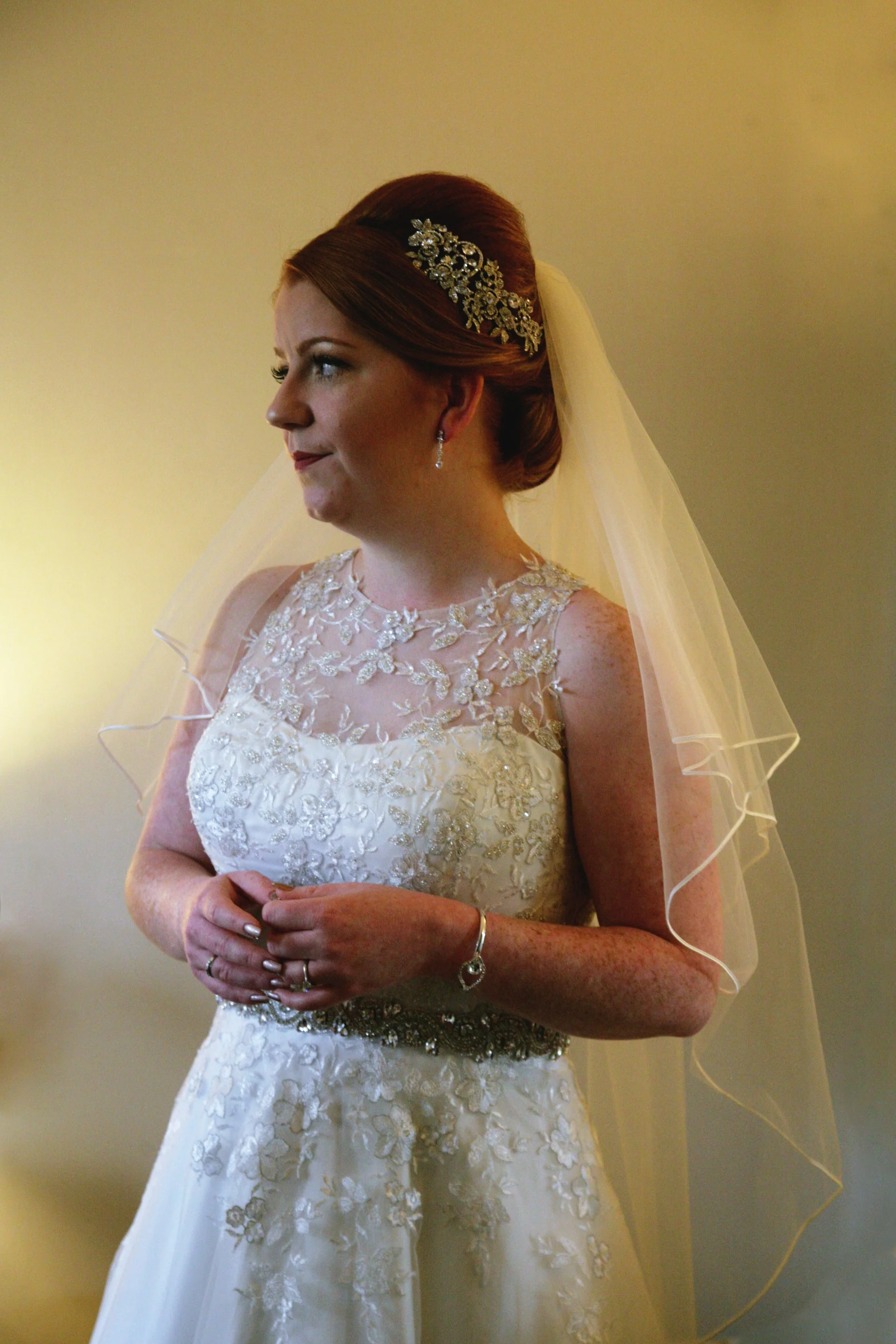 A bride in a wedding dress with lace details, wearing a veil, jewelry, and a decorative hairpiece, standing indoors.