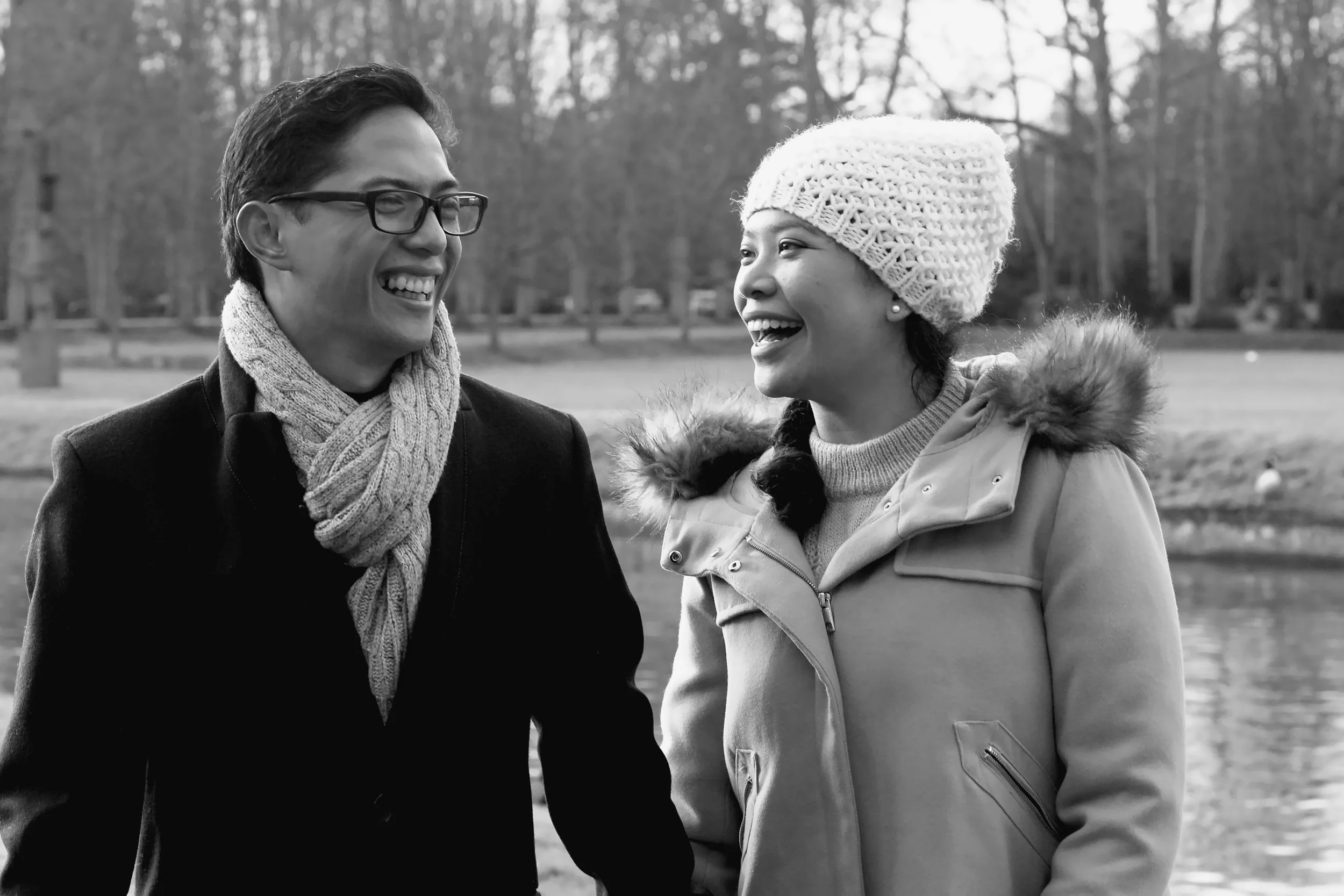 A man and a woman smiling and talking outdoors in winter, near a body of water with trees in the background.