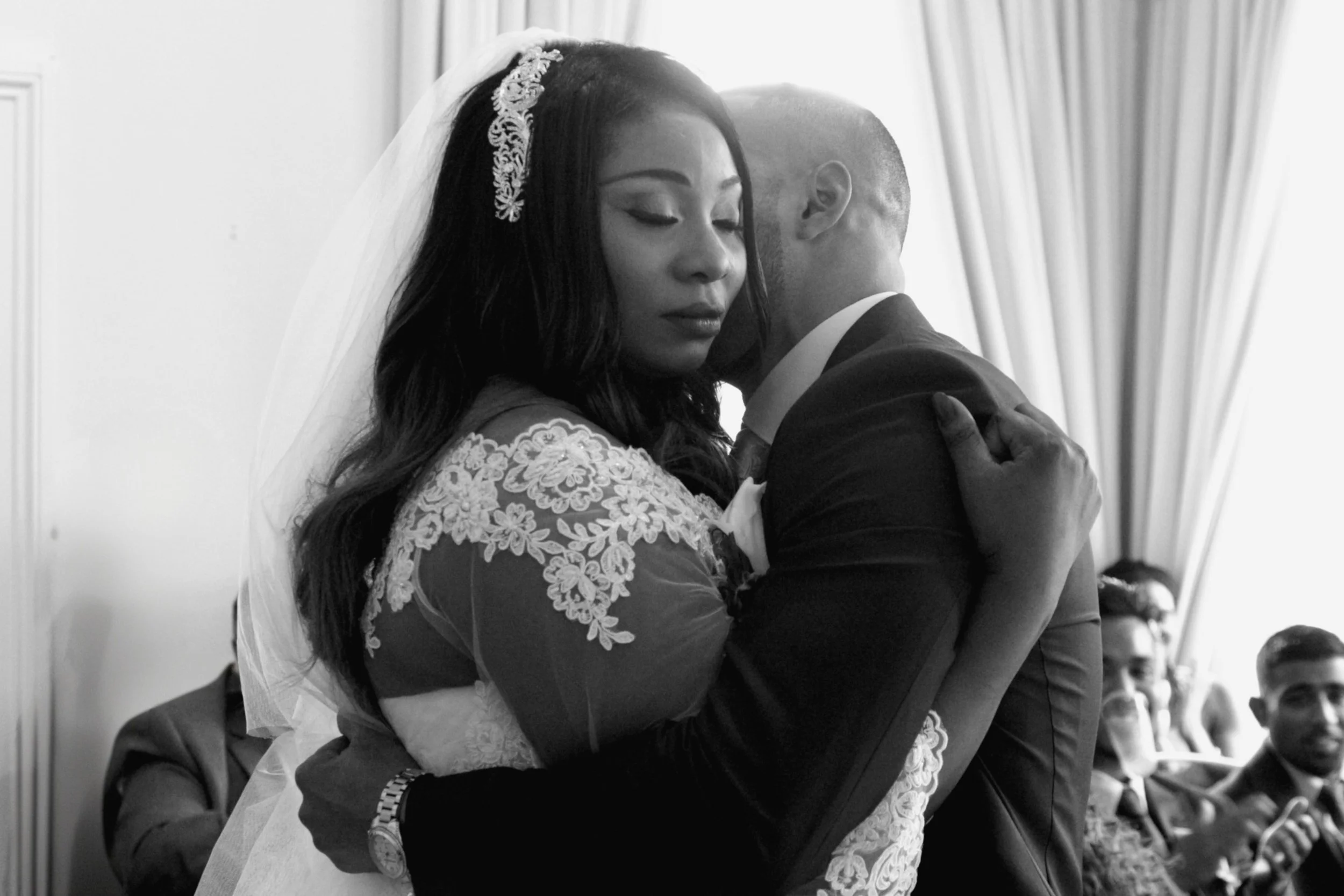 A bride and groom hugging closely during their wedding ceremony, with wedding guests visible in the background.