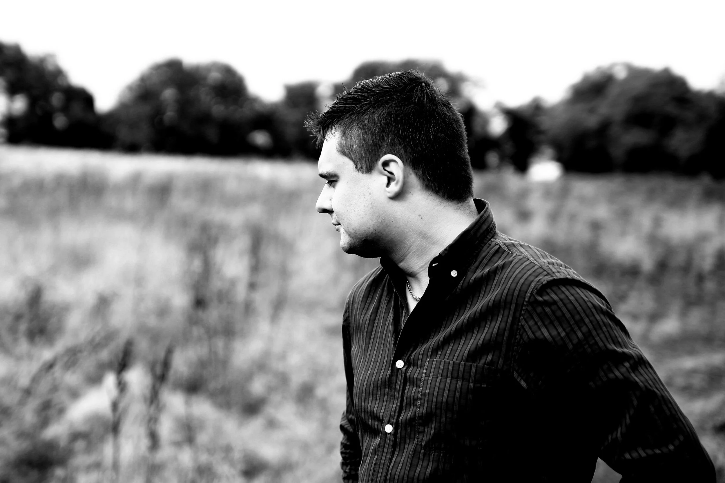A young man in profile, standing outdoors in a field with trees in the background.