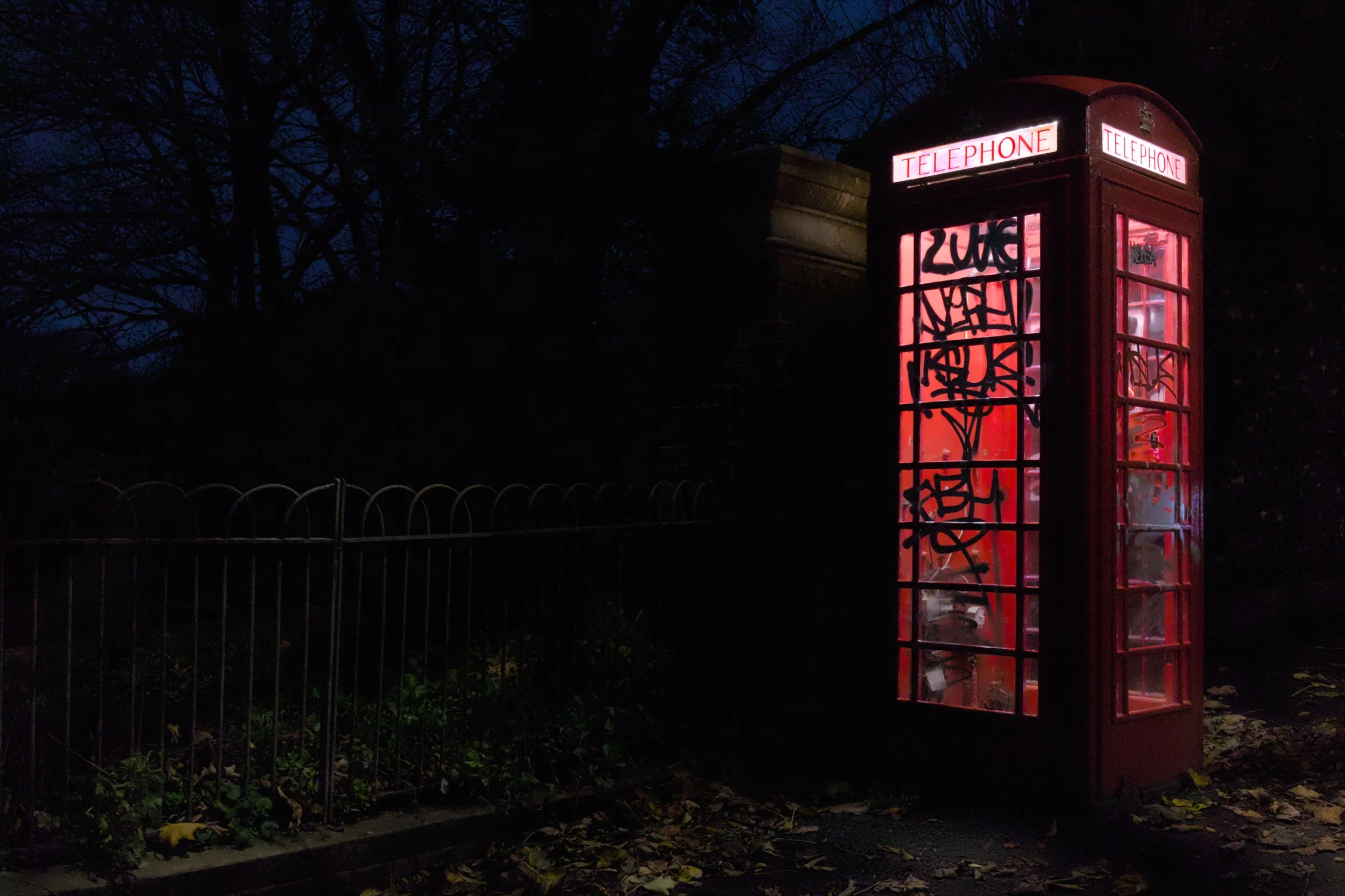 Red British telephone booth illuminated at night, with graffiti on the glass panels, next to a black metal fence with fallen leaves on the ground.