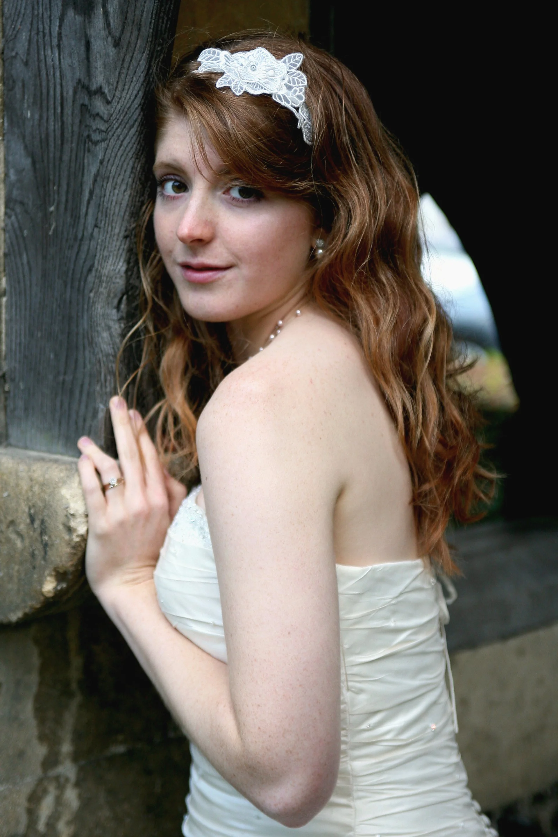 A young woman with long, curly red hair, wearing a white strapless dress, a lace headband, and jewelry, standing outdoors near a wooden structure, looking at the camera.